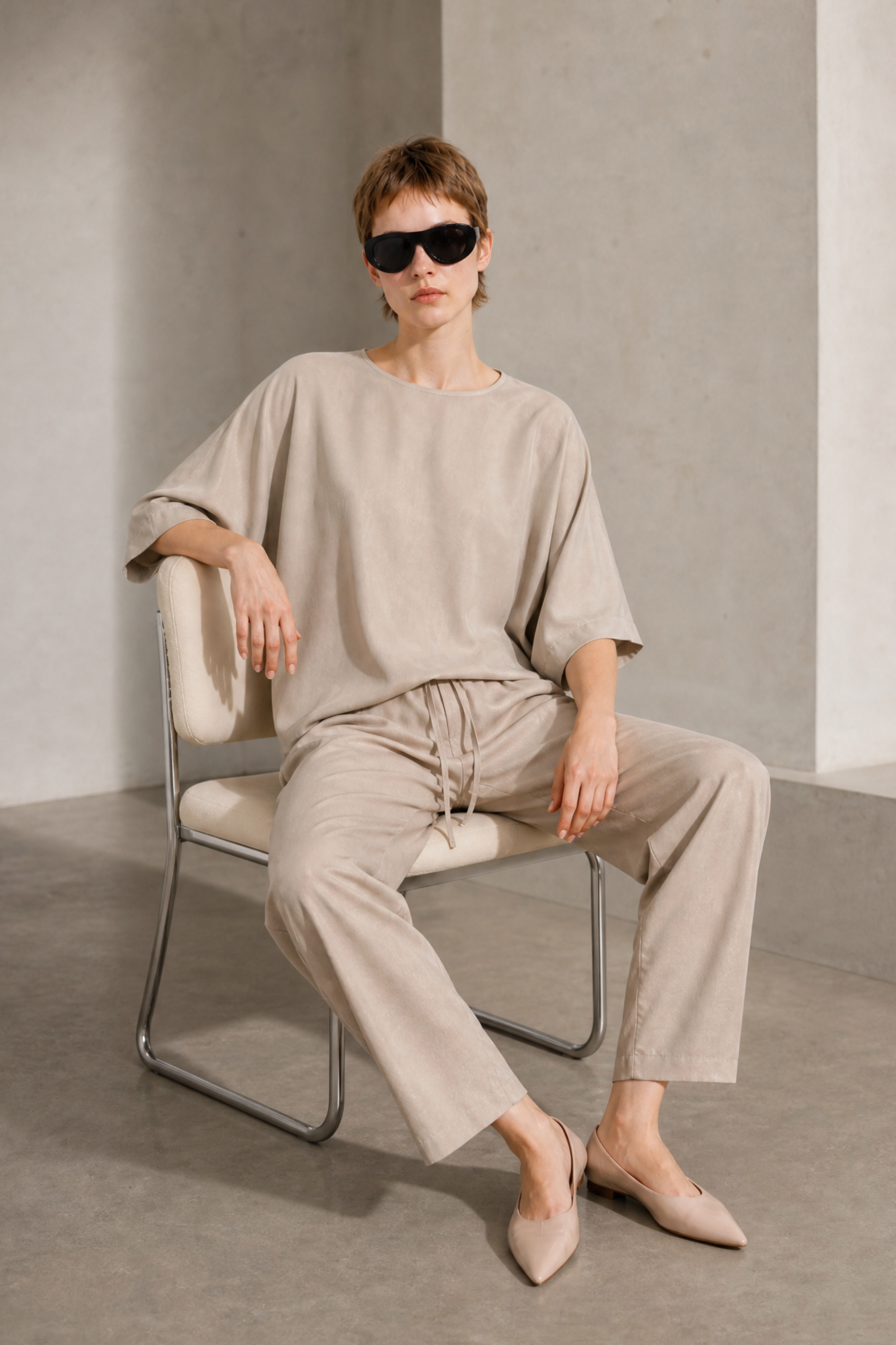 A model wearing a beige linen-blend lounge set consisting of a loose short-sleeve top and matching drawstring trousers, seated on a minimalist metal chair in a concrete-walled room.