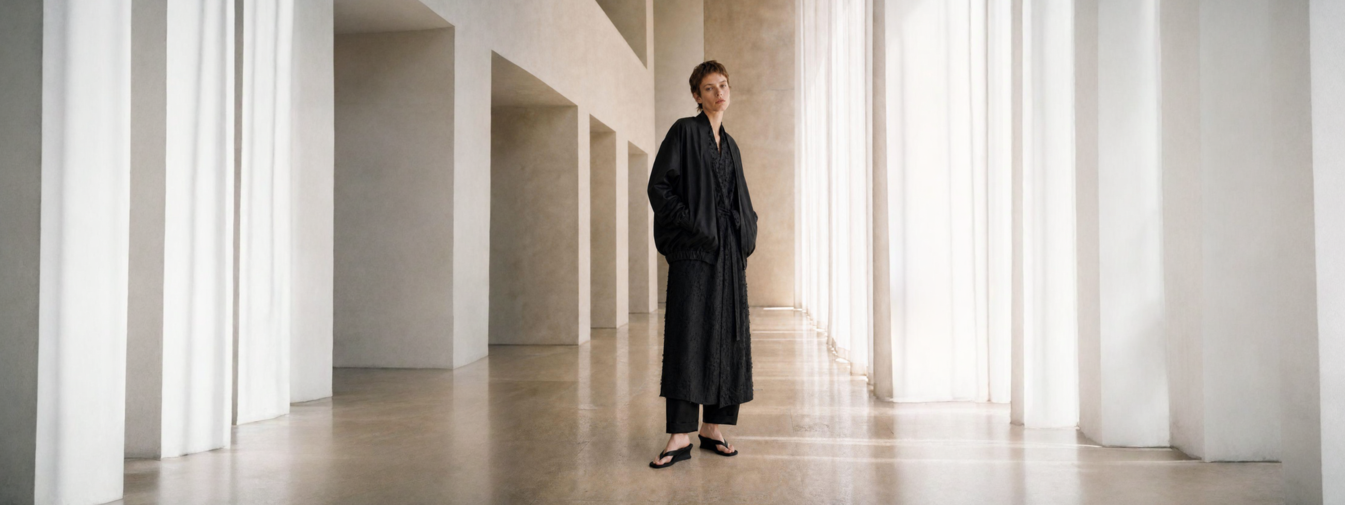 A person wearing a long black robe stands in a bright, minimalist hallway with tall white columns and soft natural light.