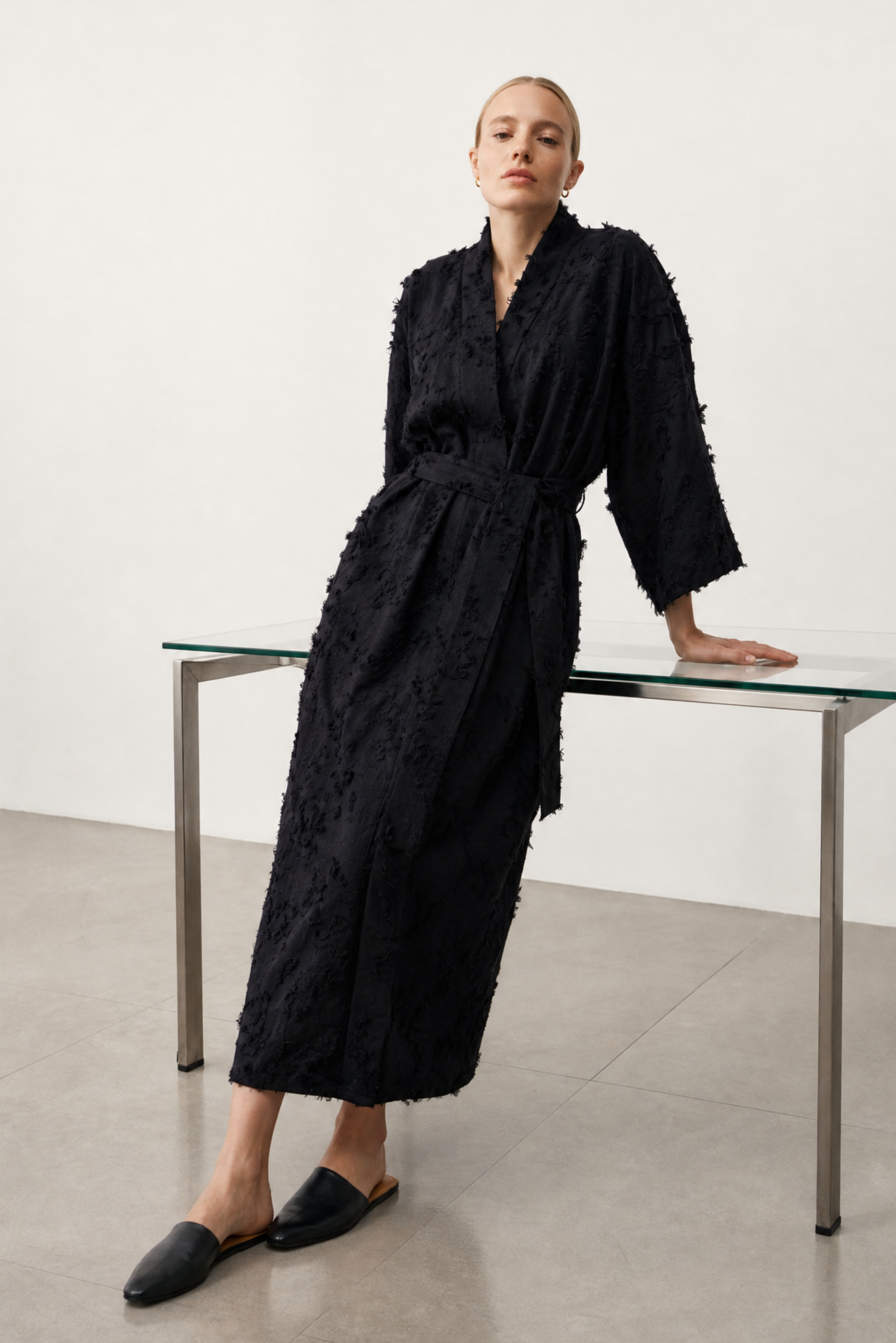 A model wearing a black textured, fringed long-sleeve robe and black mule loafers, leaning on a glass-topped table in a minimalist studio setting.
