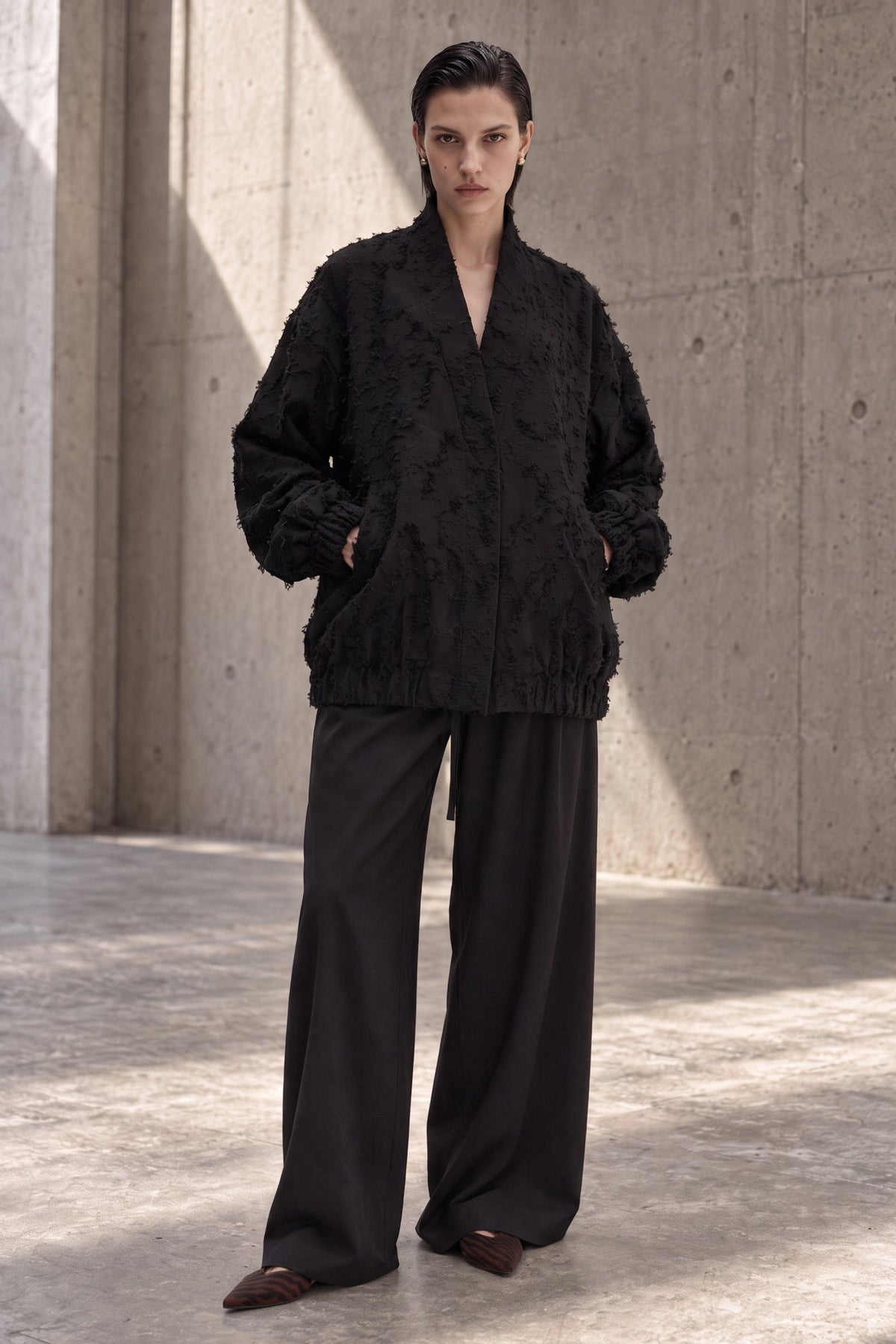 A model stands in a concrete courtyard wearing a black textured, open-front jacket with wide-leg black trousers and dark brown pointed shoes.