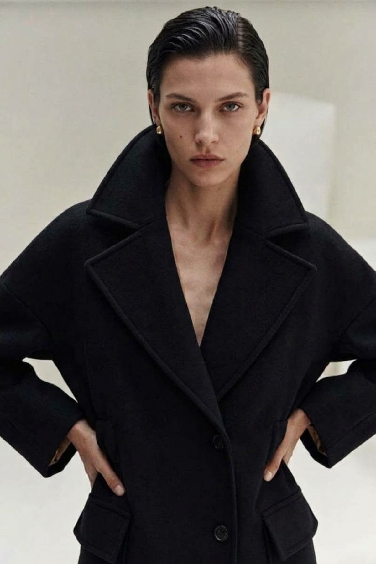 A model with slicked-back dark hair wearing a black wool-blend coat with a large collar, photographed against a neutral light gray background.