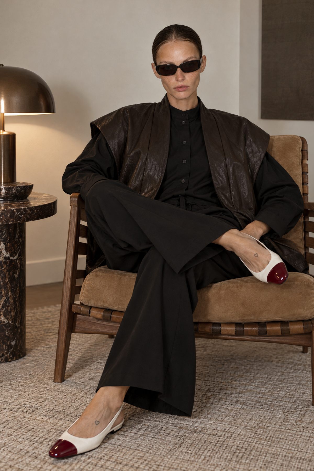 A model sits in a mid-century modern armchair wearing a black outfit with a brown leather vest, black sunglasses, and white pointed-toe flats with burgundy caps, in a styled room with a side table and lamp.