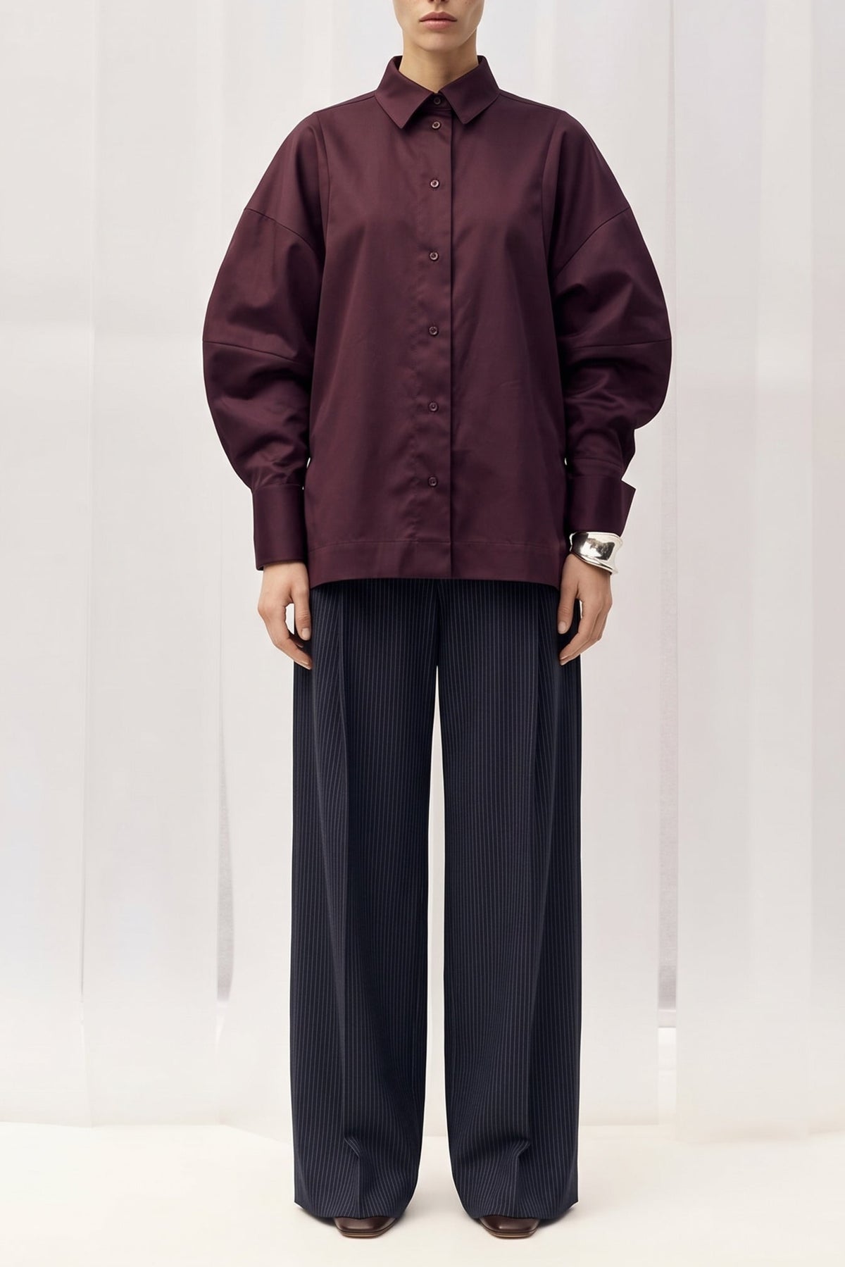 A model stands facing forward wearing an oversized long-sleeve button-up shirt in deep plum purple and dark wide-leg trousers, set against a white curtain backdrop.