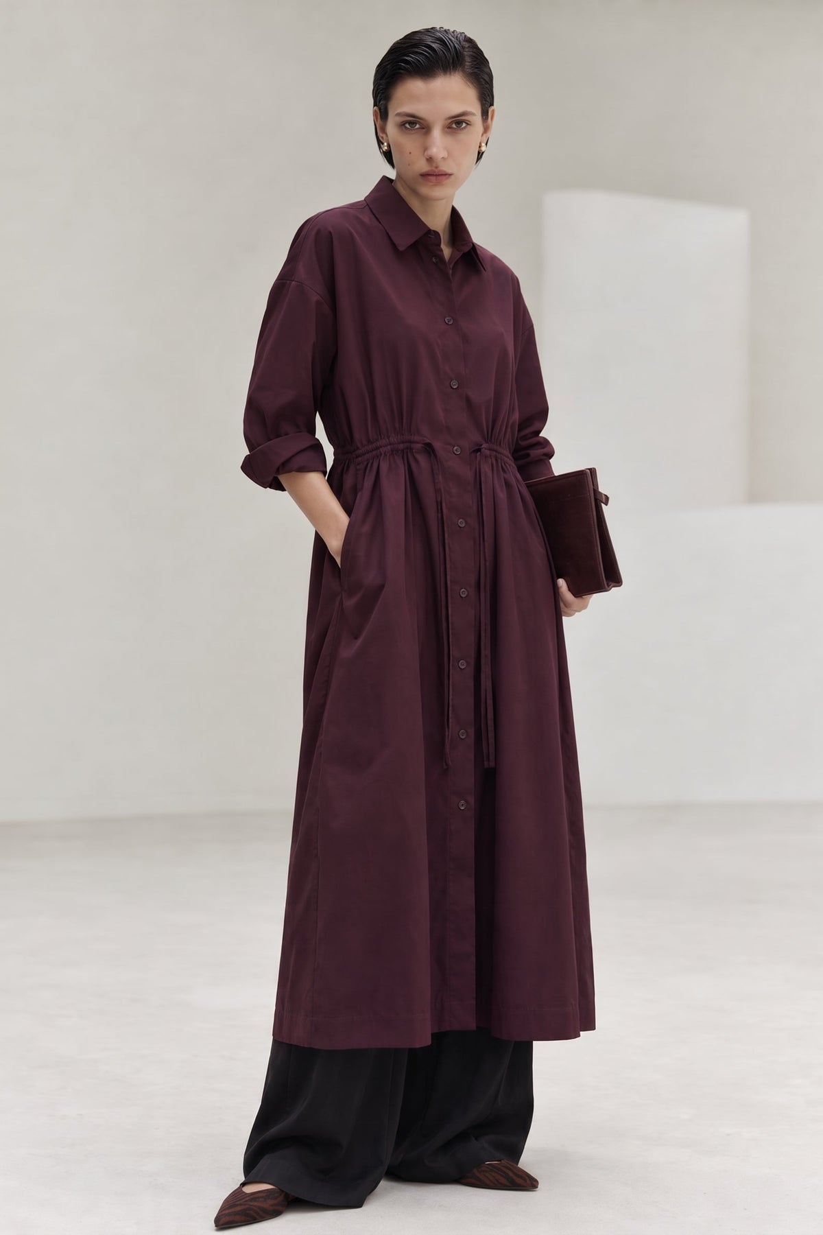 A model stands in a minimalist studio wearing a long, dark plum button-front shirt dress with a tie waist belt, paired with black wide-leg pants and holding a matching dark clutch.
