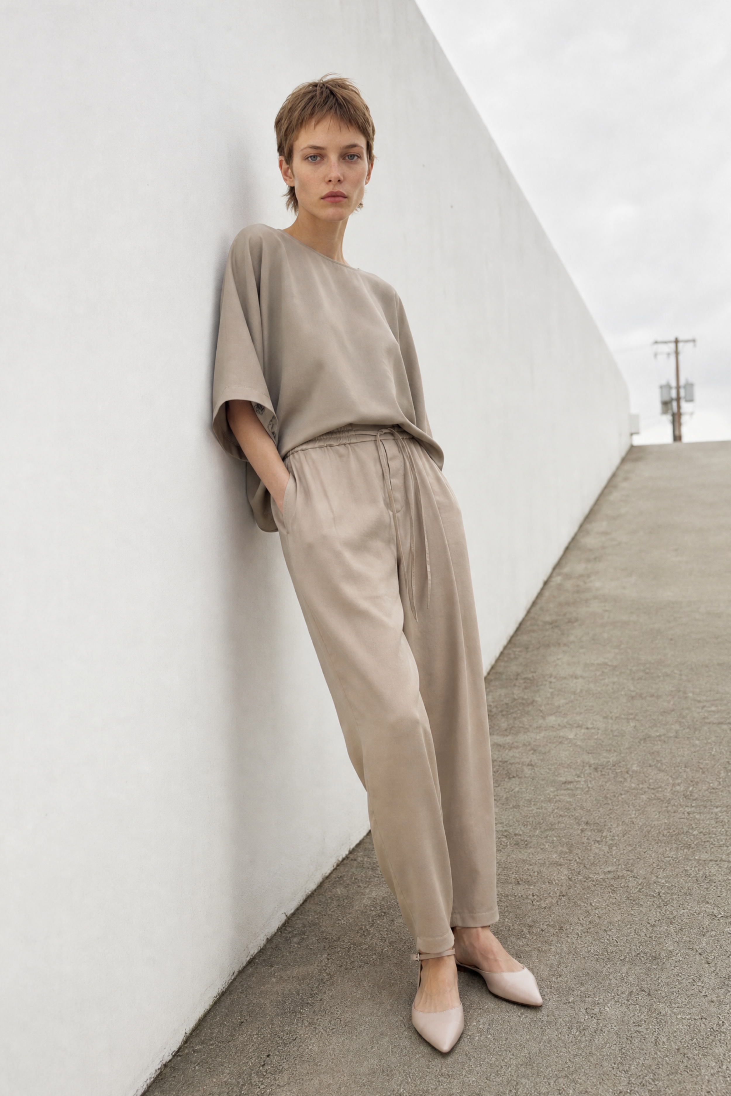 A woman stands against a white concrete wall wearing a loose taupe short-sleeve top and matching beige wide-leg trousers, paired with light beige pointed flats.