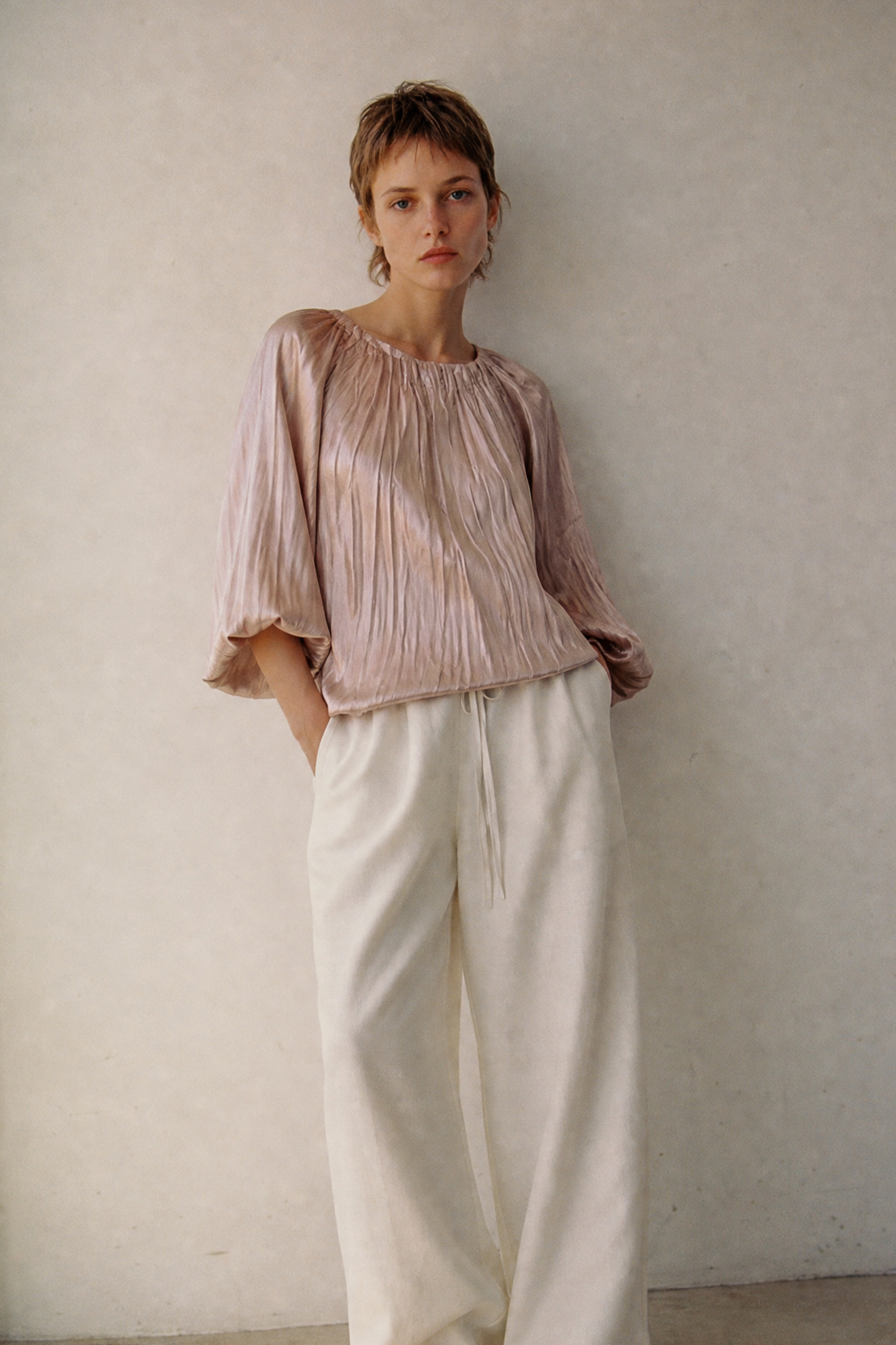 Woman wearing a pink blouse and white pants against a plain wall.