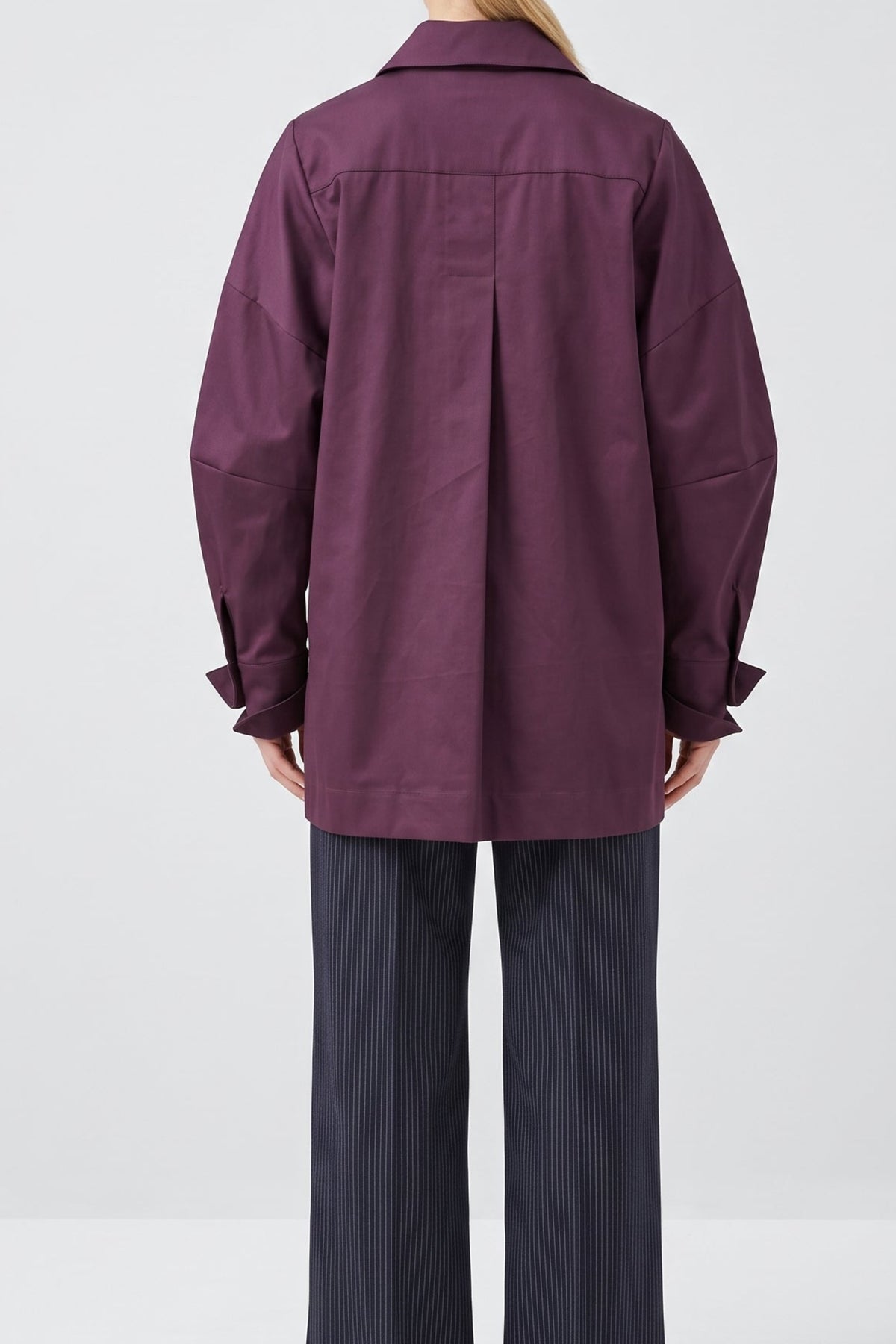 A woman viewed from the back wearing a loose, oversized plum-purple button-up shirt and dark charcoal pinstripe trousers, standing against a light gray studio background.