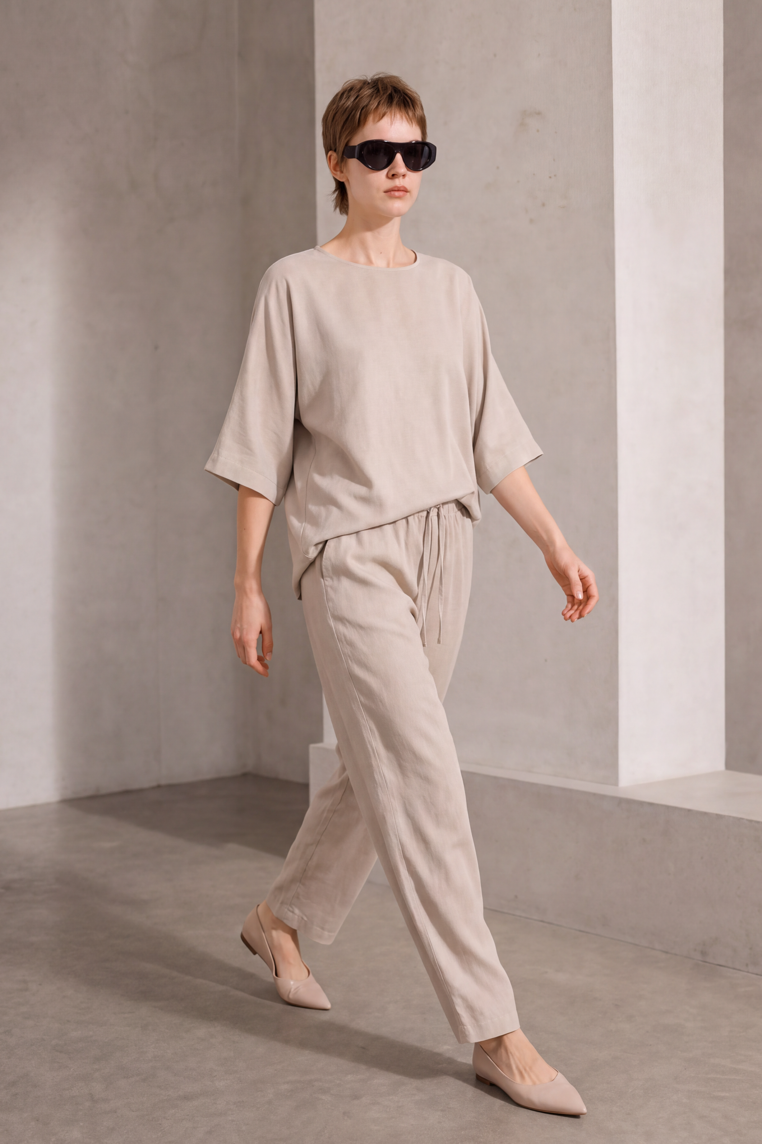 A model wearing a light beige linen two-piece set consisting of a short-sleeve top and matching drawstring trousers, styled with beige pointed-toe flats and black sunglasses, in a minimalist concrete room.