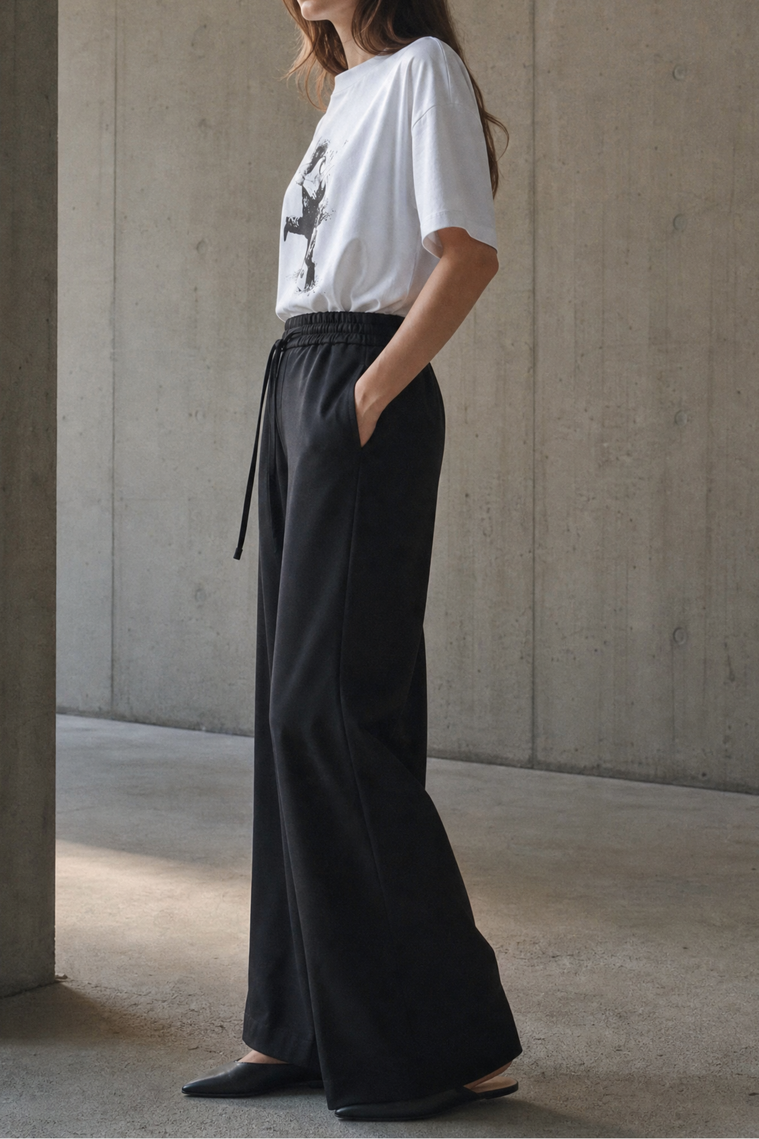 Woman wearing a white t-shirt and black wide-leg pants standing against a concrete wall.