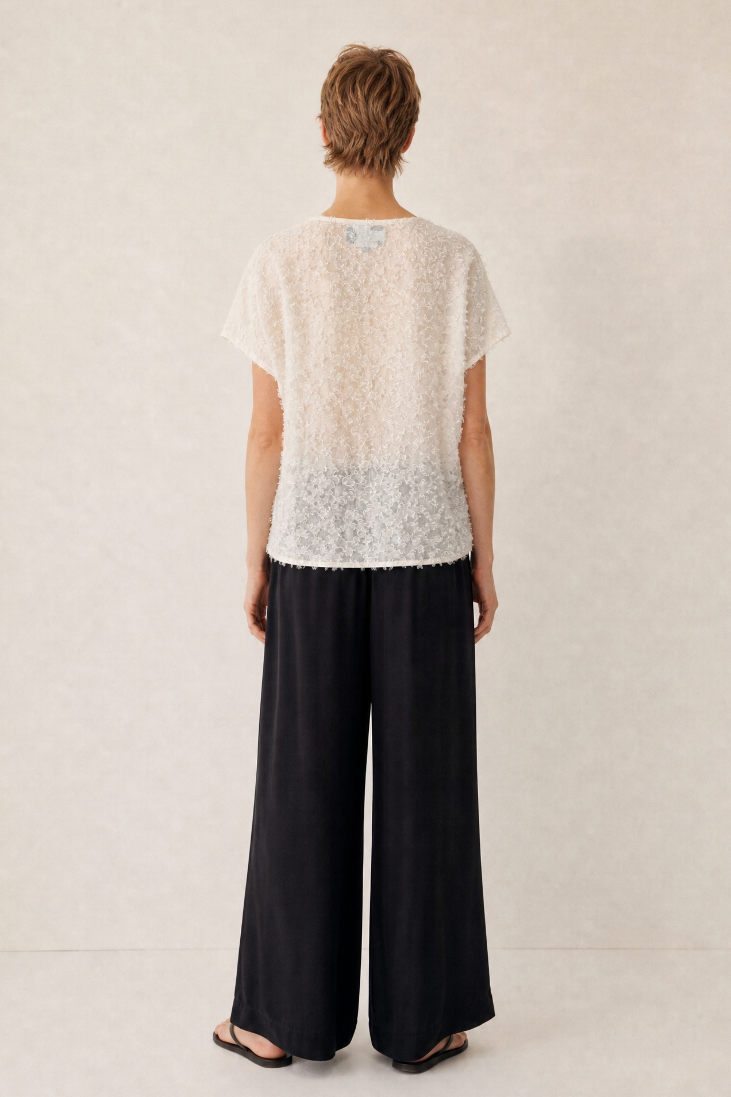 A model shown from the back wearing a short-sleeve white lace top and black wide-leg trousers, standing against a neutral textured beige background.