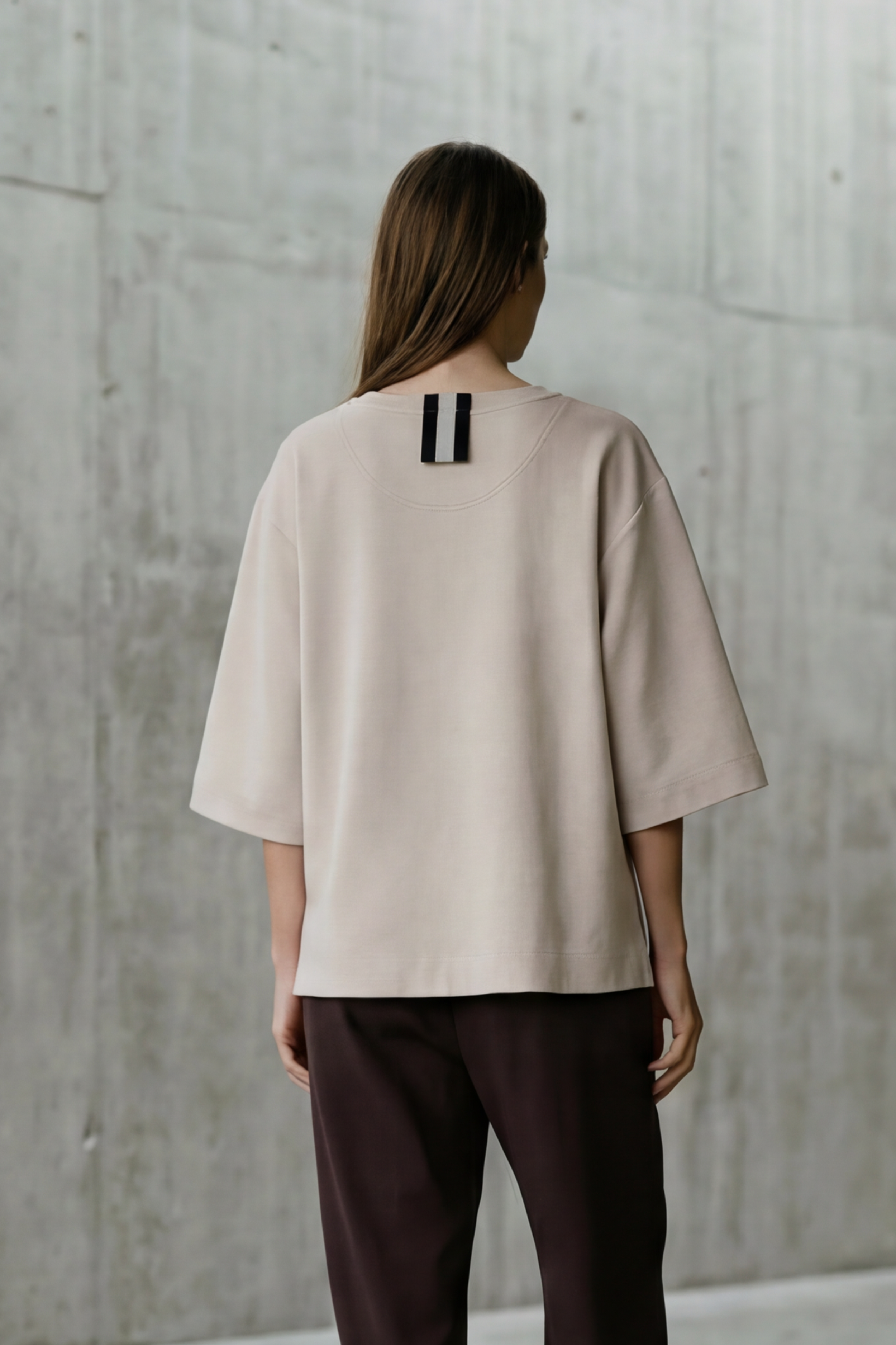 Person wearing a beige shirt and dark pants standing in a minimalist concrete room.