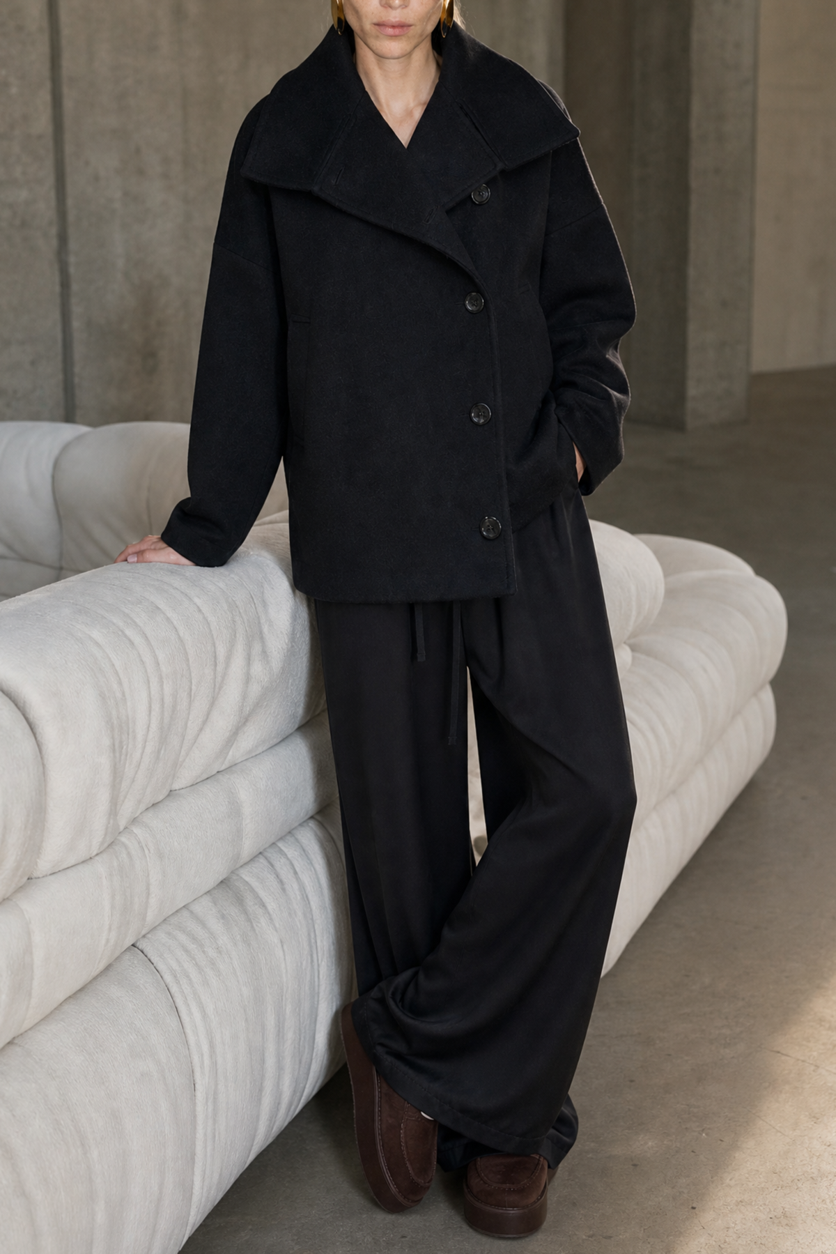 A model stands in a minimalist concrete room next to a white curved sofa, wearing a black double-breasted coat and matching wide-leg trousers with brown shoes.