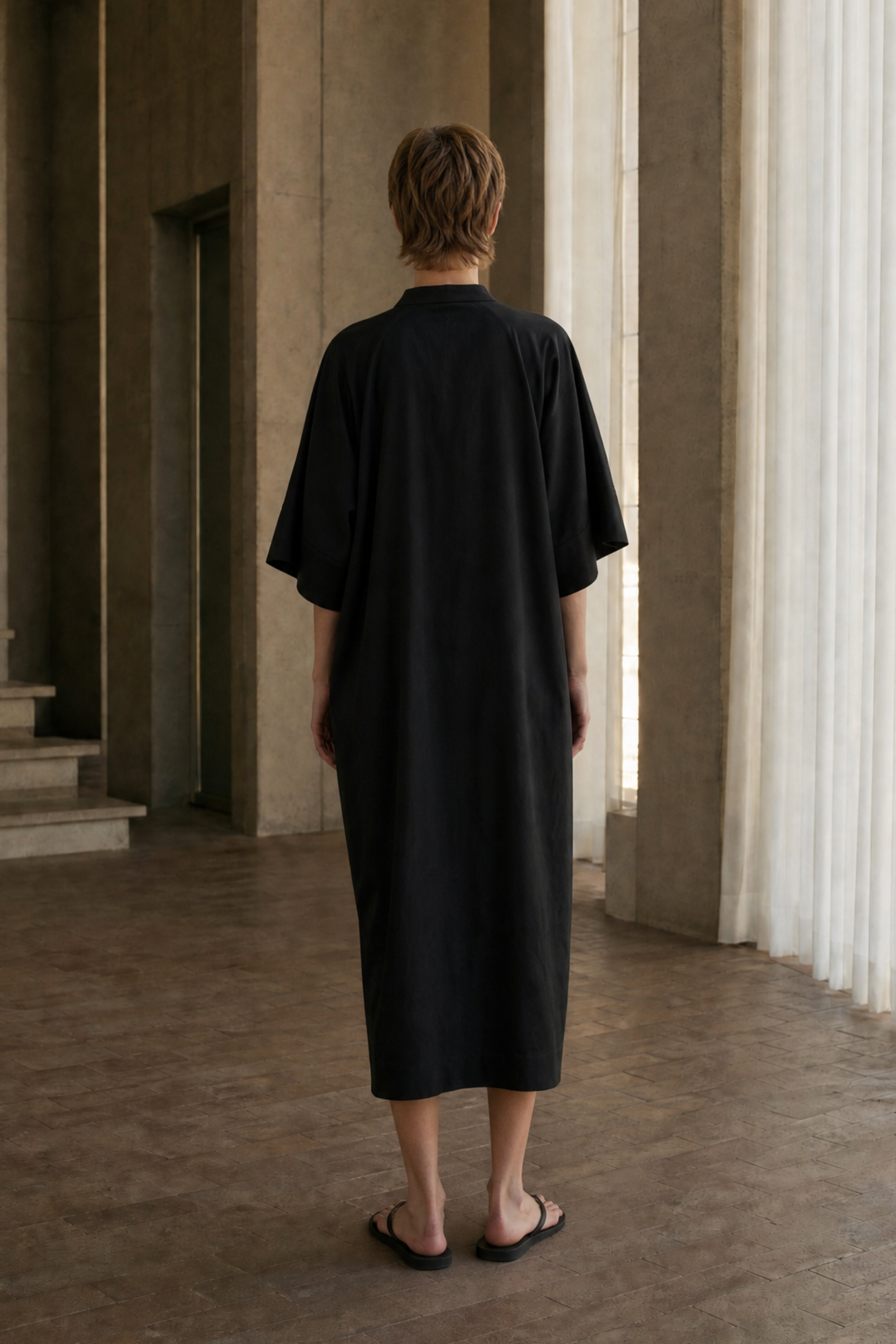 A person viewed from behind wearing a loose black midi dress or kimono-style tunic in a minimalist concrete interior with large windows and curtains.