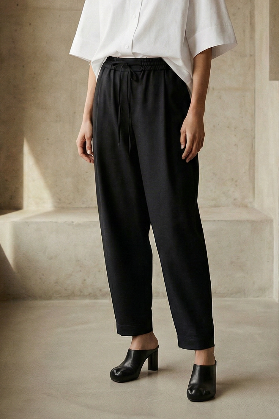 Woman wearing a white blouse and black pants in a minimalistic room with concrete walls and floor.