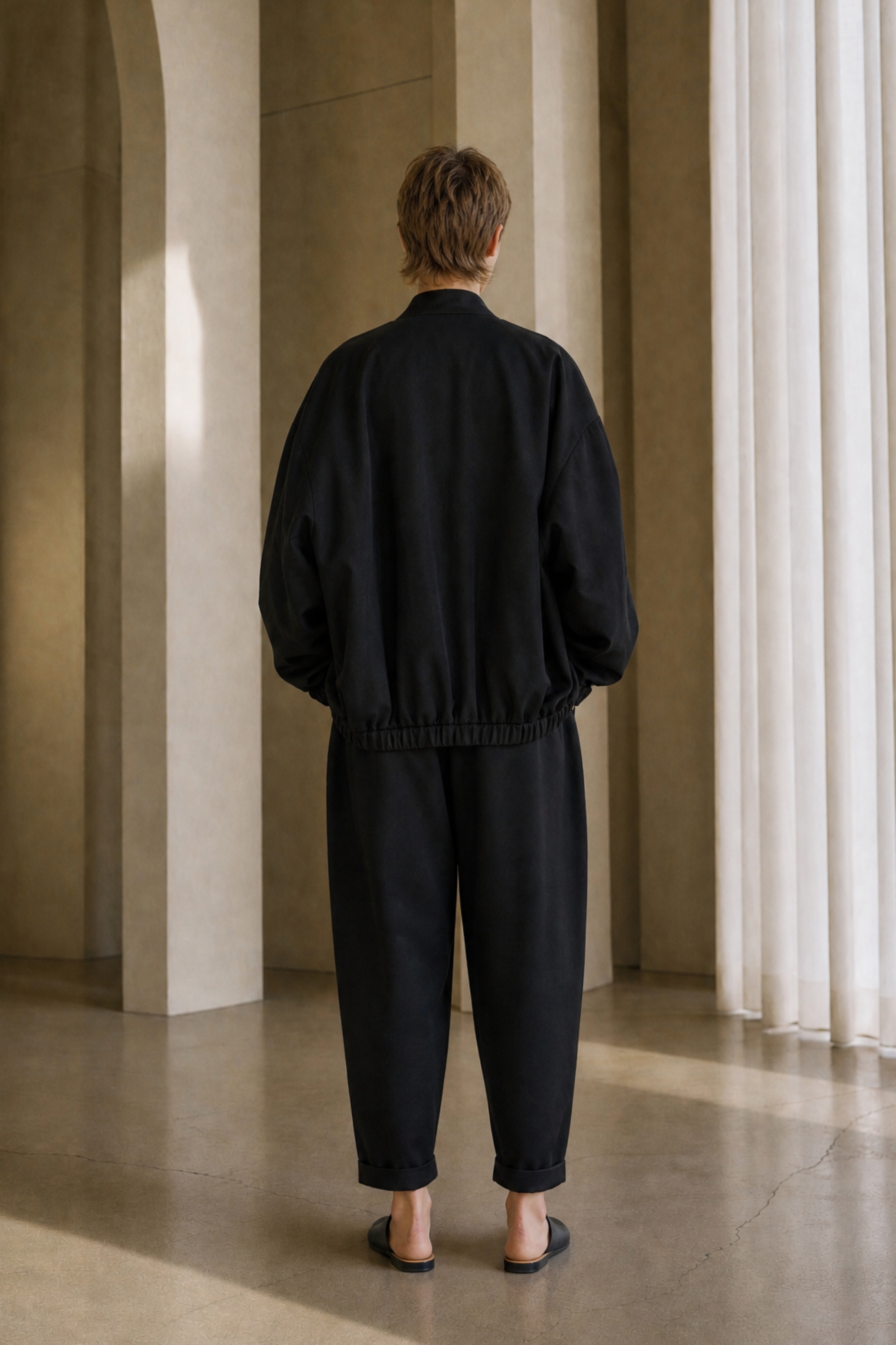 A person viewed from behind wearing a black bomber jacket and black wide-leg pants, standing in a minimalist interior with beige walls, arches, and white sheer curtains.