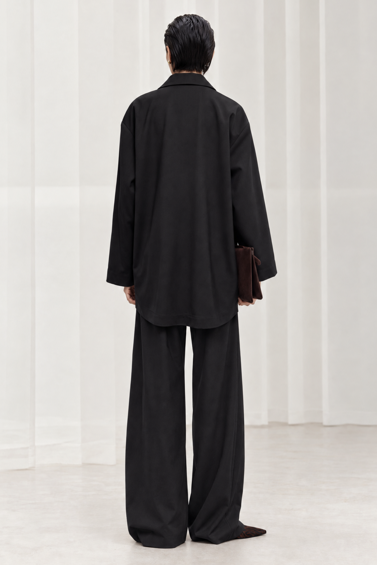 A model viewed from behind wearing an oversized black long-sleeve shirt and matching wide-leg trousers, holding a dark brown leather clutch against a minimalist white draped background.