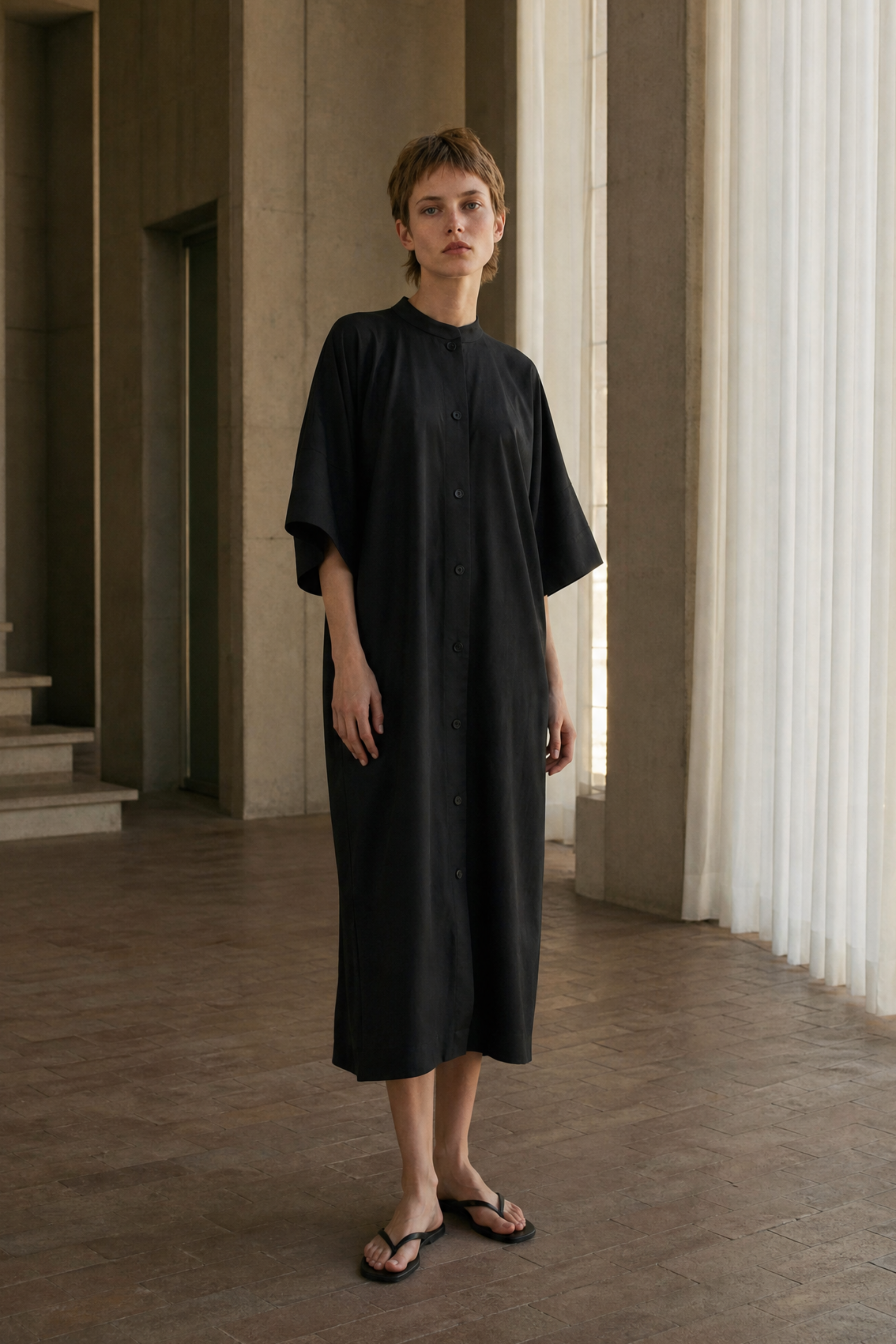 A model stands in a minimalist, modern interior with concrete columns and sheer curtains, wearing a loose-fitting black midi dress and black sandals.