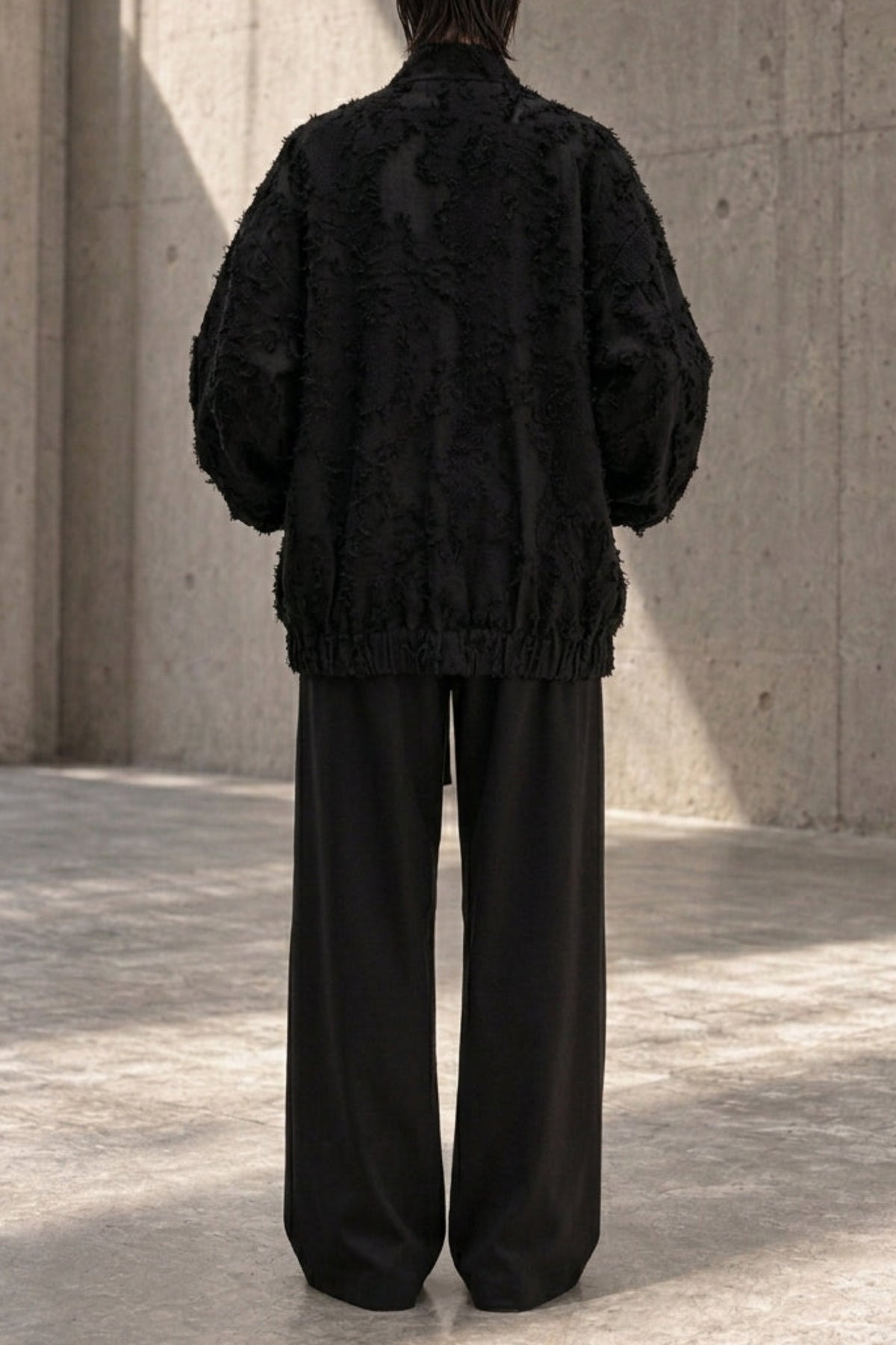 A model viewed from behind wearing a black textured, voluminous jacket and black wide-leg trousers, standing in front of a concrete wall.