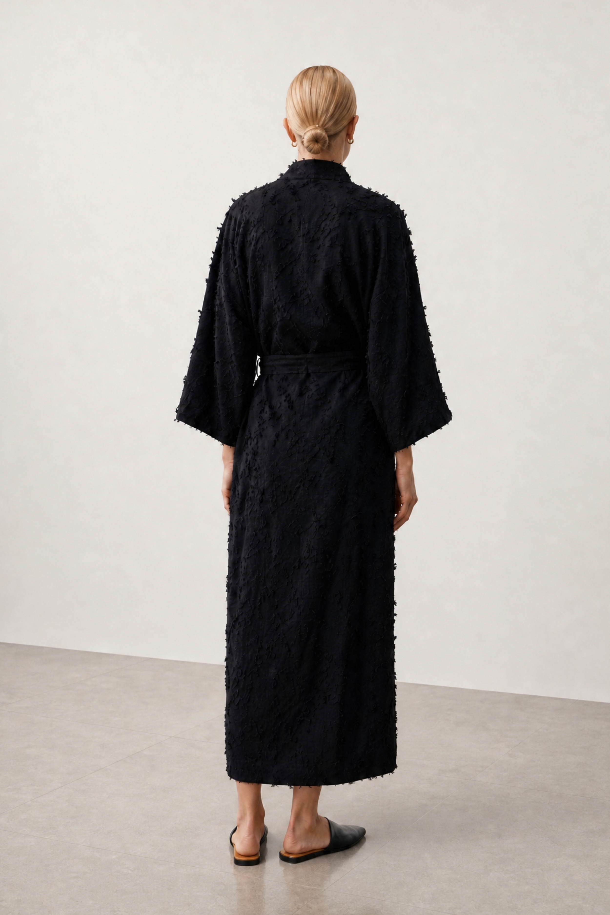 A model viewed from behind wearing a long black textured robe with wide sleeves and a tie belt, standing in a minimalist studio with a light gray wall and concrete floor.