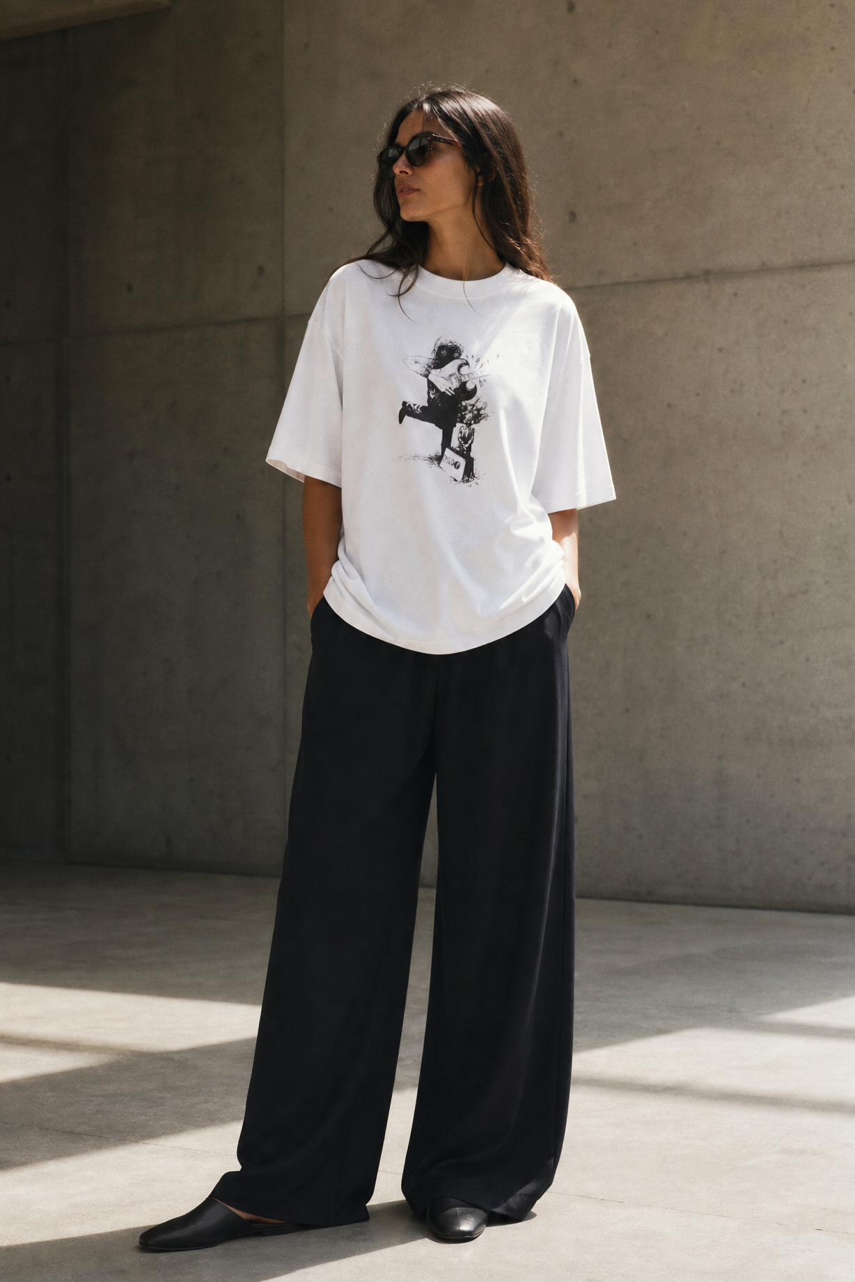 Person wearing a white t-shirt with a graphic design and black pants against a concrete wall.