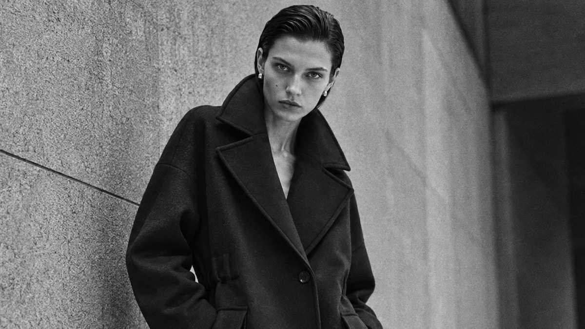 A model wearing a long black coat stands against a textured concrete wall in a black-and-white urban setting.