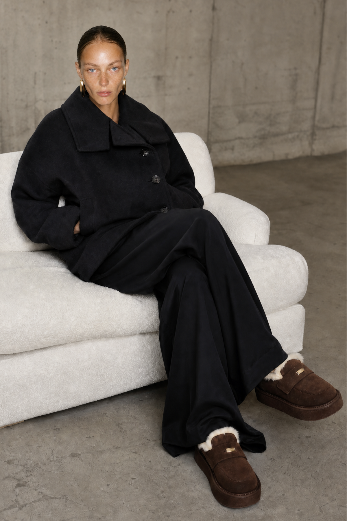 A model sits on a white boucle sofa wearing a black wool-blend coat and wide-leg trousers, paired with brown suede slip-on slippers with white shearling lining, set against a concrete wall and floor.