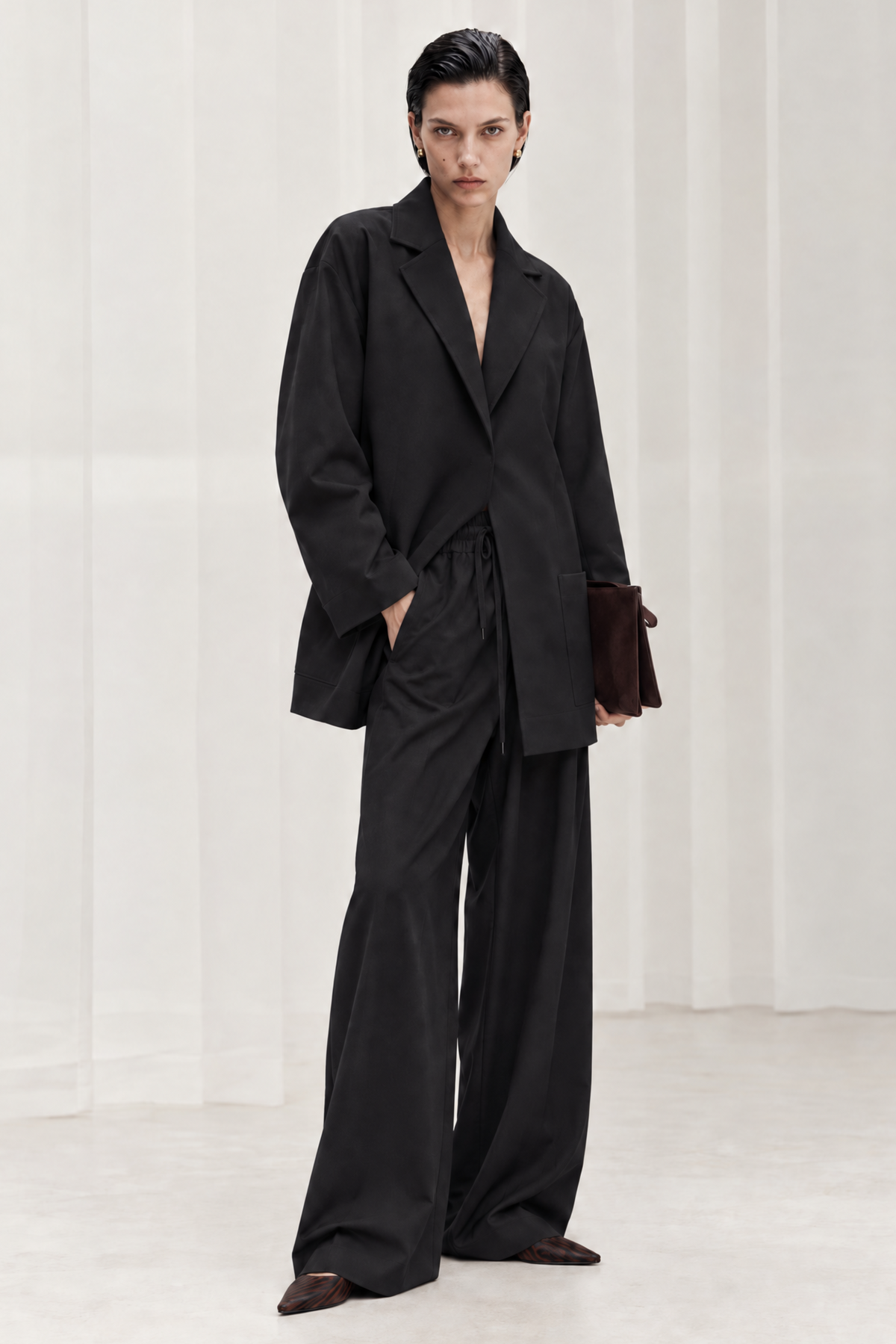 A model stands in a minimalist studio wearing a black oversized blazer and matching wide-leg trousers, holding a dark brown leather clutch.