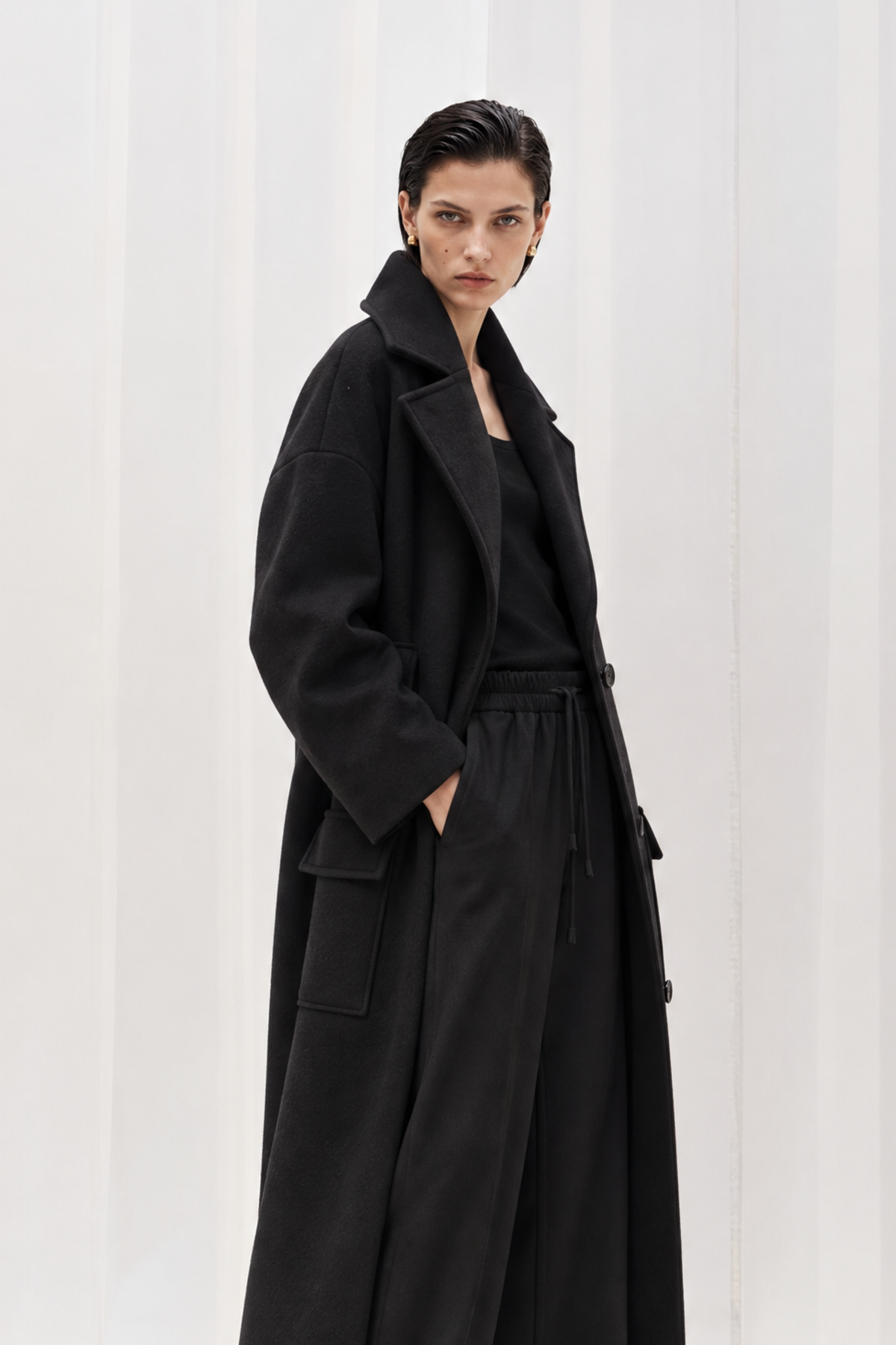 A model wearing a long black coat with a tie waist, wide-leg trousers, and brown shoes, standing against a white curtain backdrop.