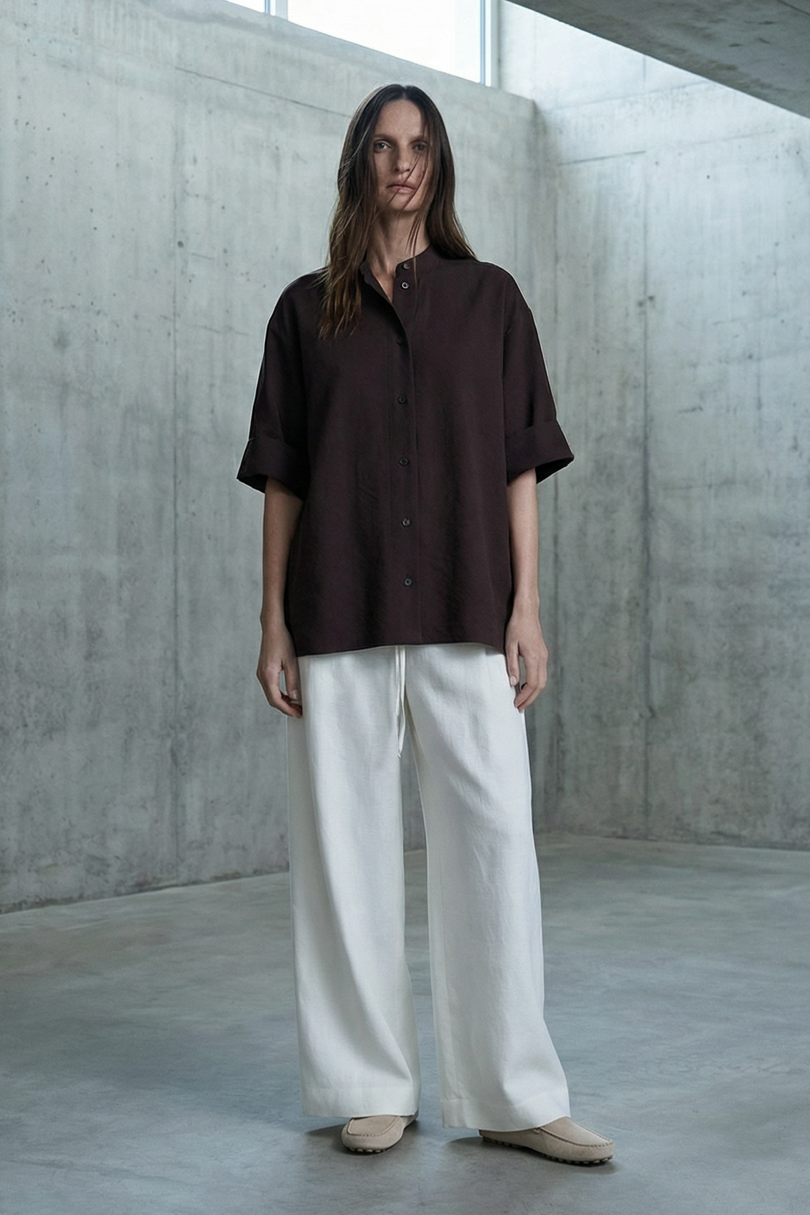 Person wearing a brown shirt and white pants standing in a concrete room.
