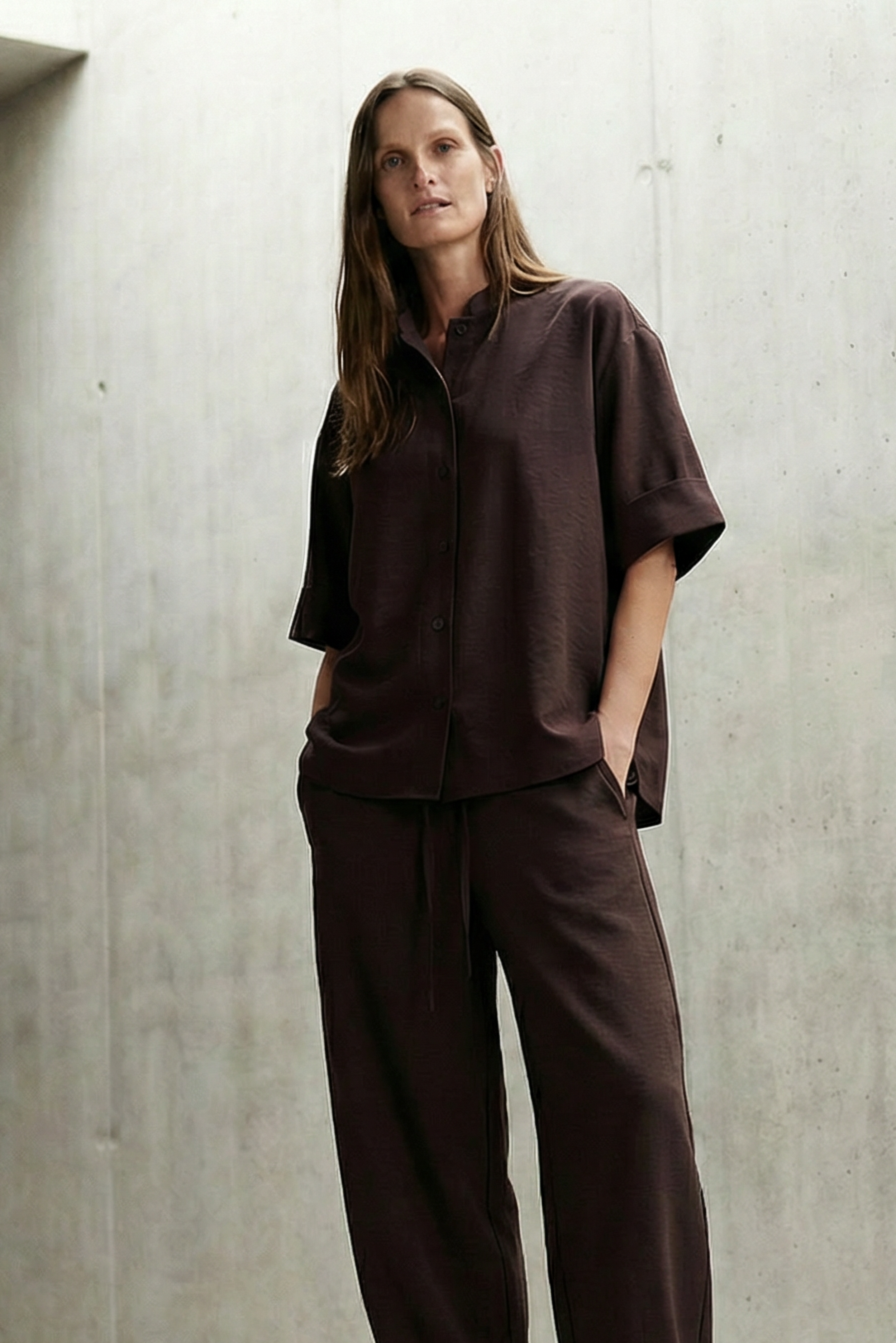 Person wearing a brown outfit standing in a minimalistic concrete room.