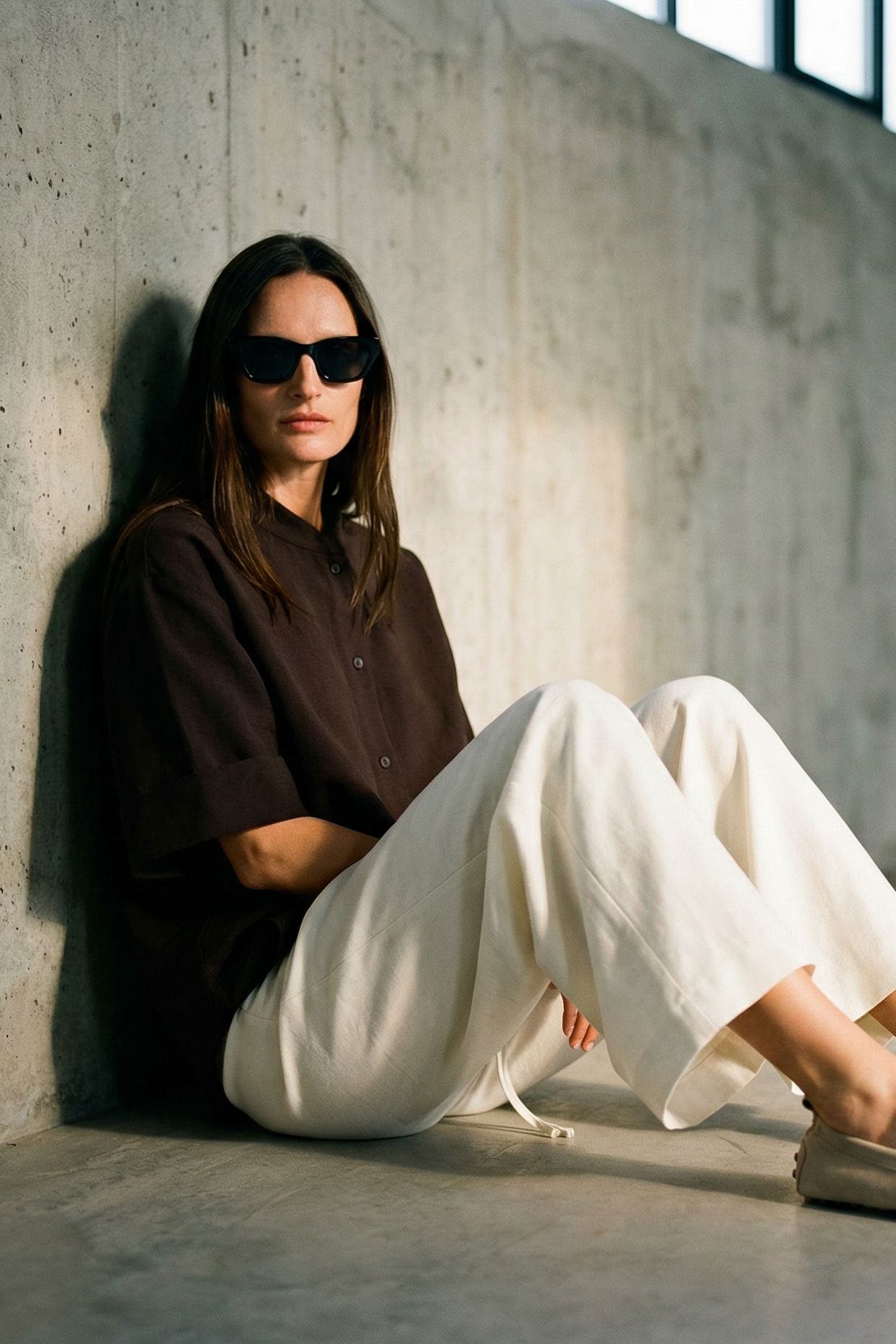 Person sitting against a concrete wall wearing sunglasses and casual clothing.