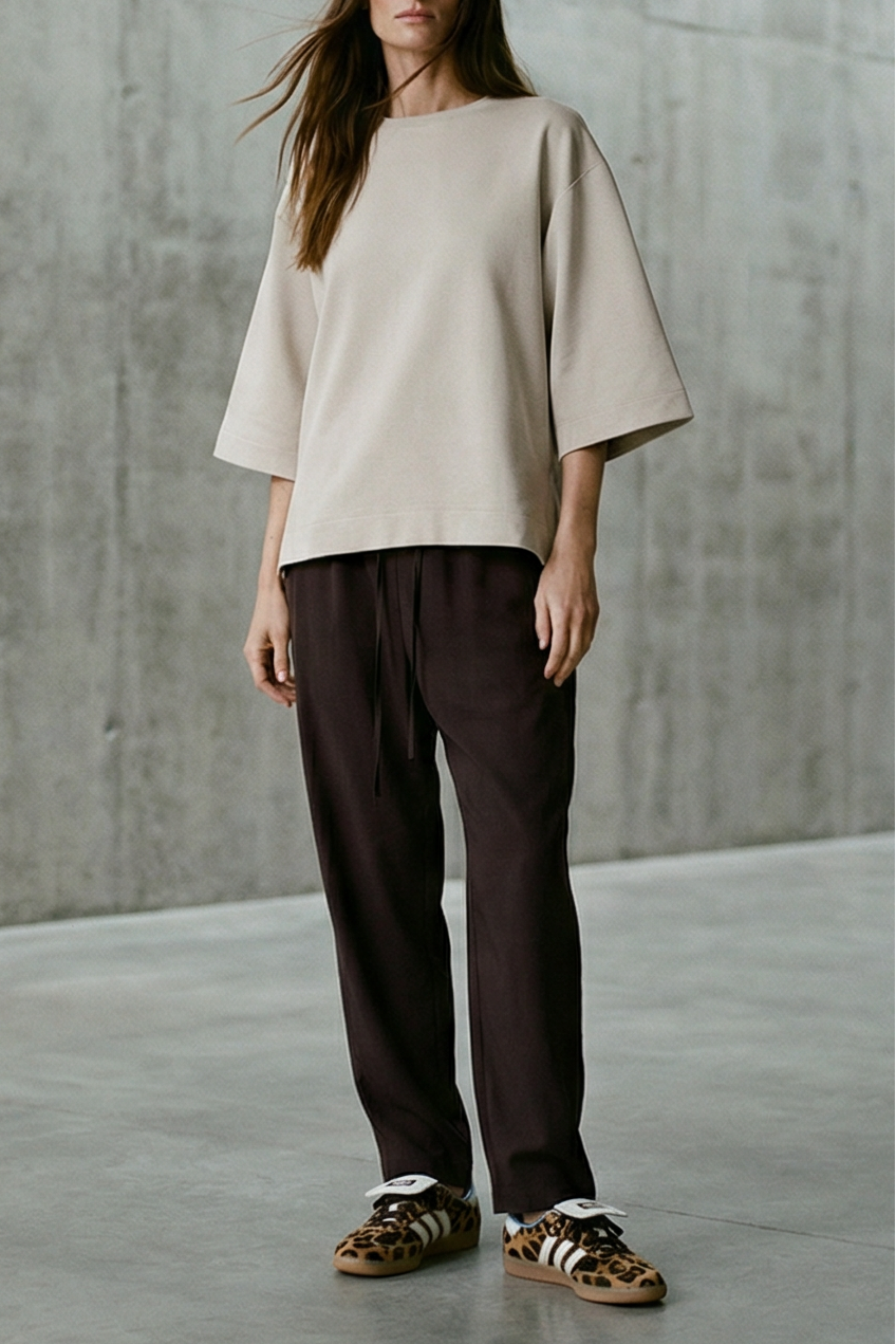 Woman wearing a beige top and dark pants standing in a minimalist room with concrete walls.
