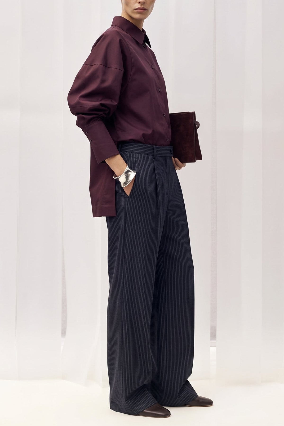 A model stands in profile wearing a dark burgundy long-sleeve button-up shirt with voluminous balloon sleeves, paired with dark navy wide-leg trousers and holding a matching dark burgundy clutch against a white draped studio backdrop.