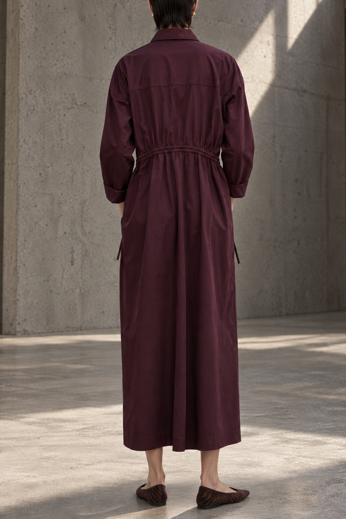 A person viewed from behind wearing a long burgundy shirt dress in a minimalist concrete building with natural light shadows.