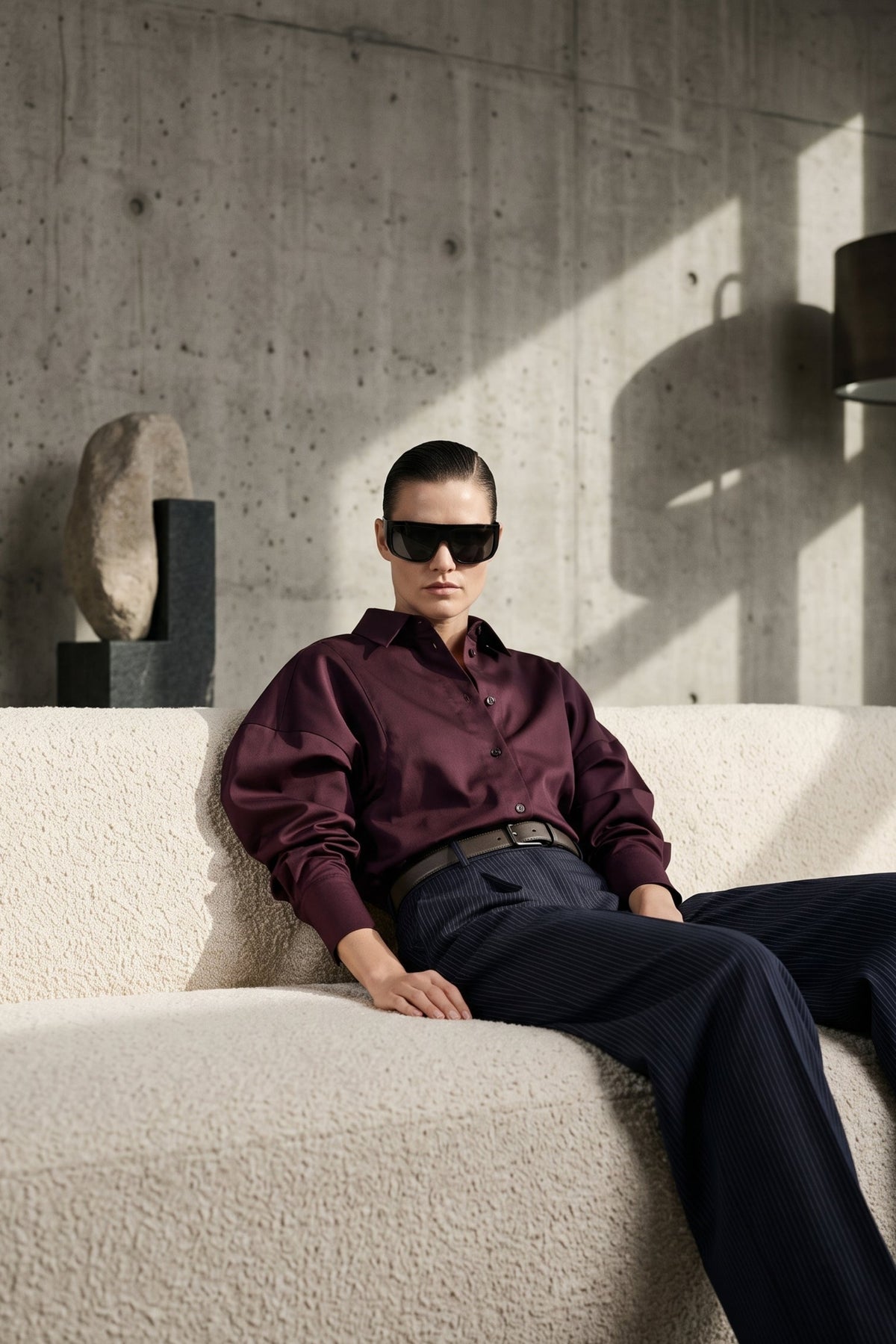A model wearing a burgundy long-sleeve button-down shirt, navy trousers, and black sunglasses sits on a textured cream boucle sofa against a concrete wall.