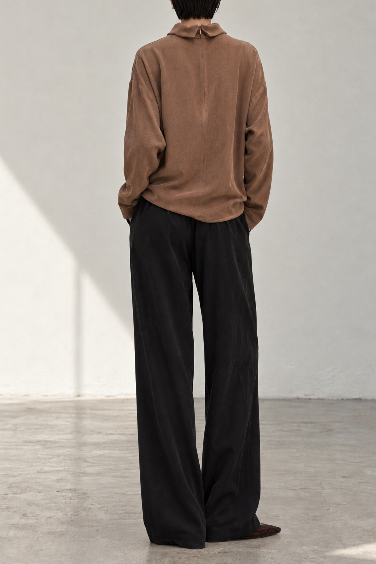 A person standing with their back to the camera, wearing a brown long-sleeve collared shirt and black wide-leg trousers in a minimalist studio setting.