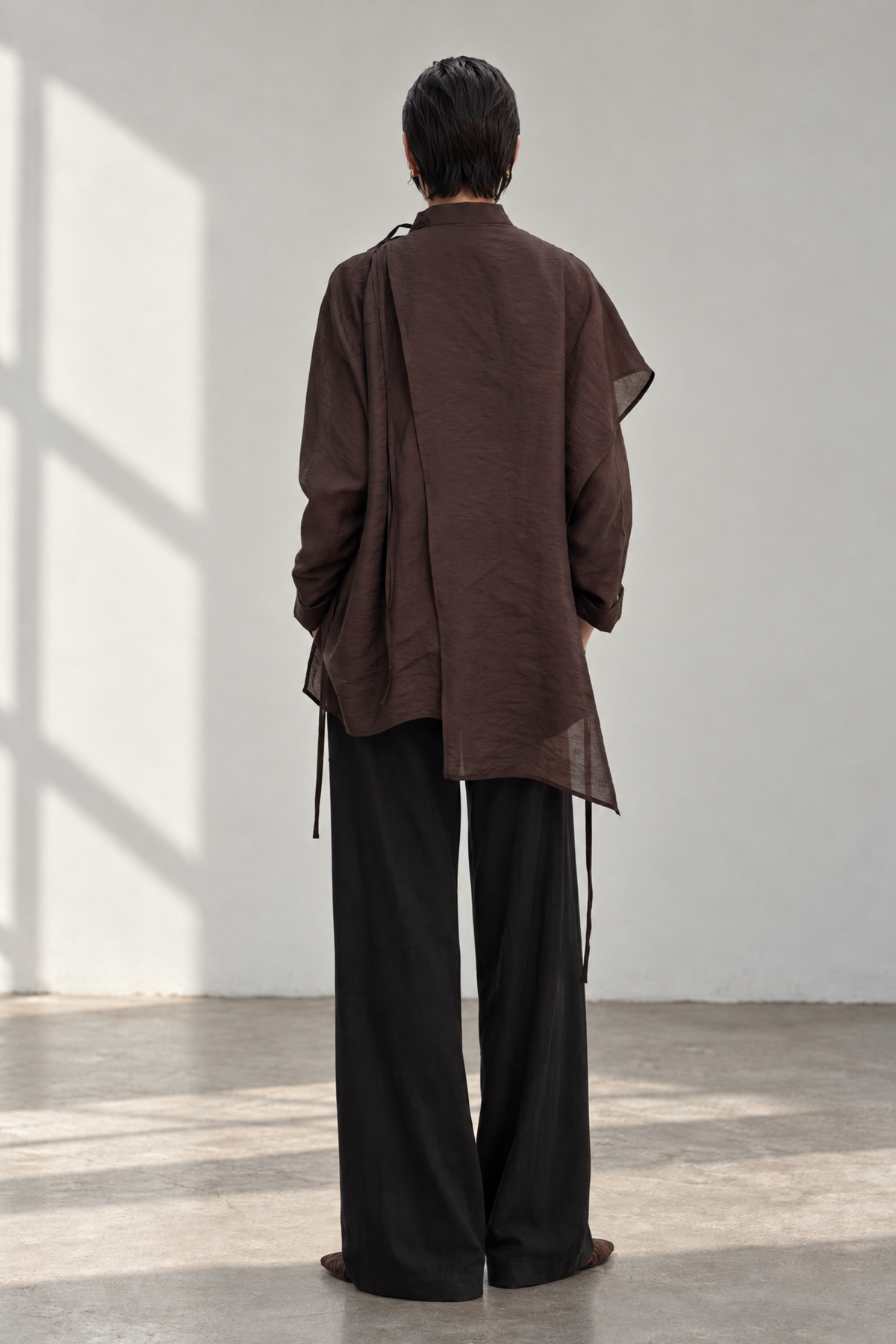 A person standing with their back to the camera, wearing a dark brown loose-fitting long-sleeve top and wide-leg black pants in a minimalist room with soft window shadows.
