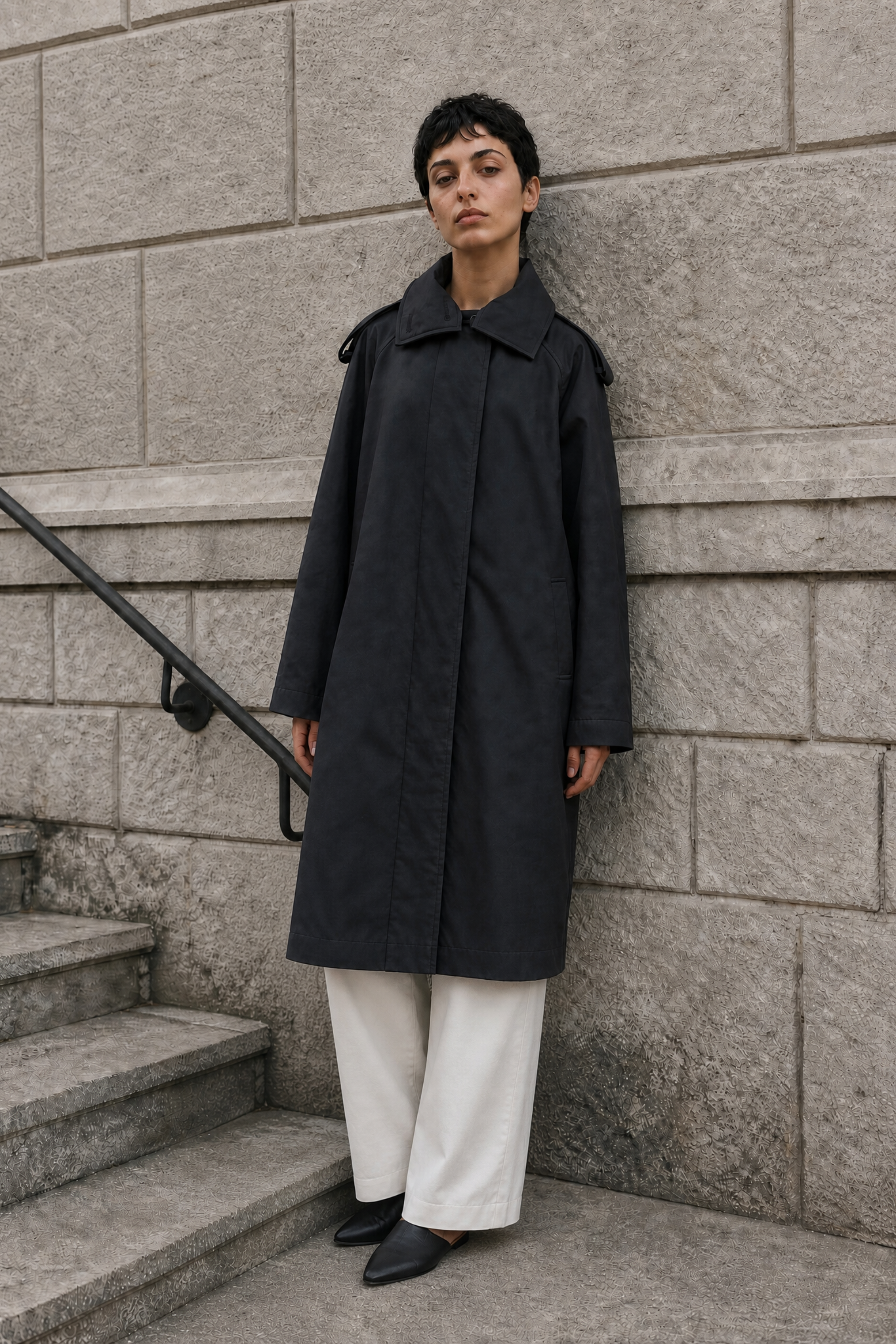 Person wearing a black coat standing on stone steps against a gray stone wall.