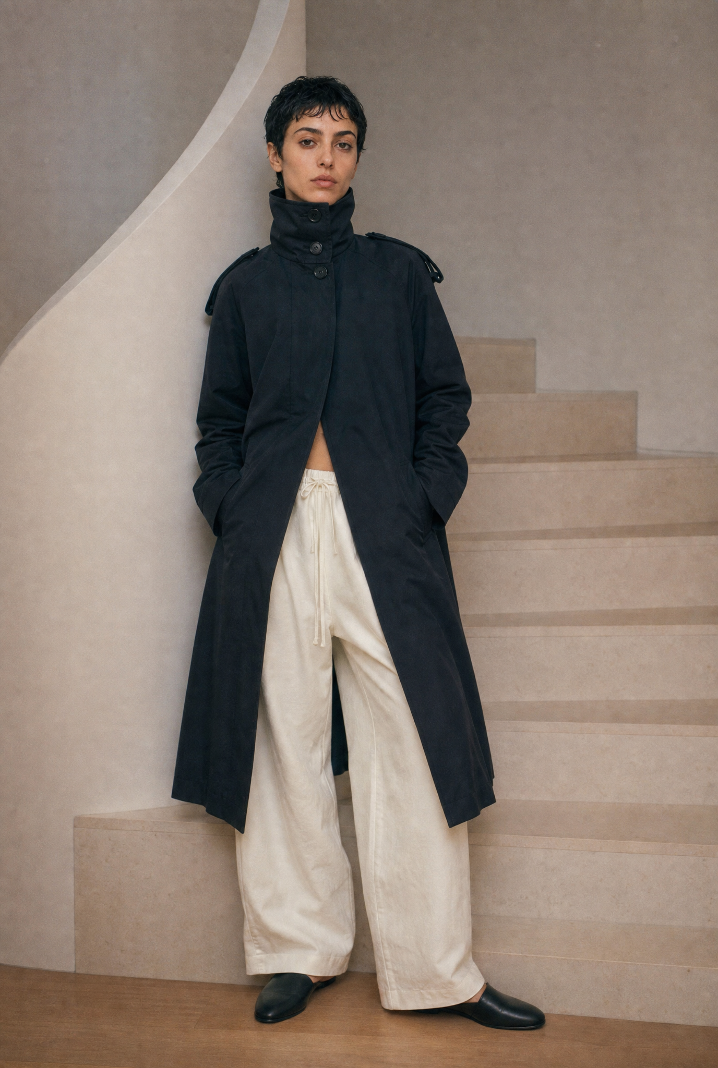 Person wearing a long dark coat and light pants standing on a staircase.