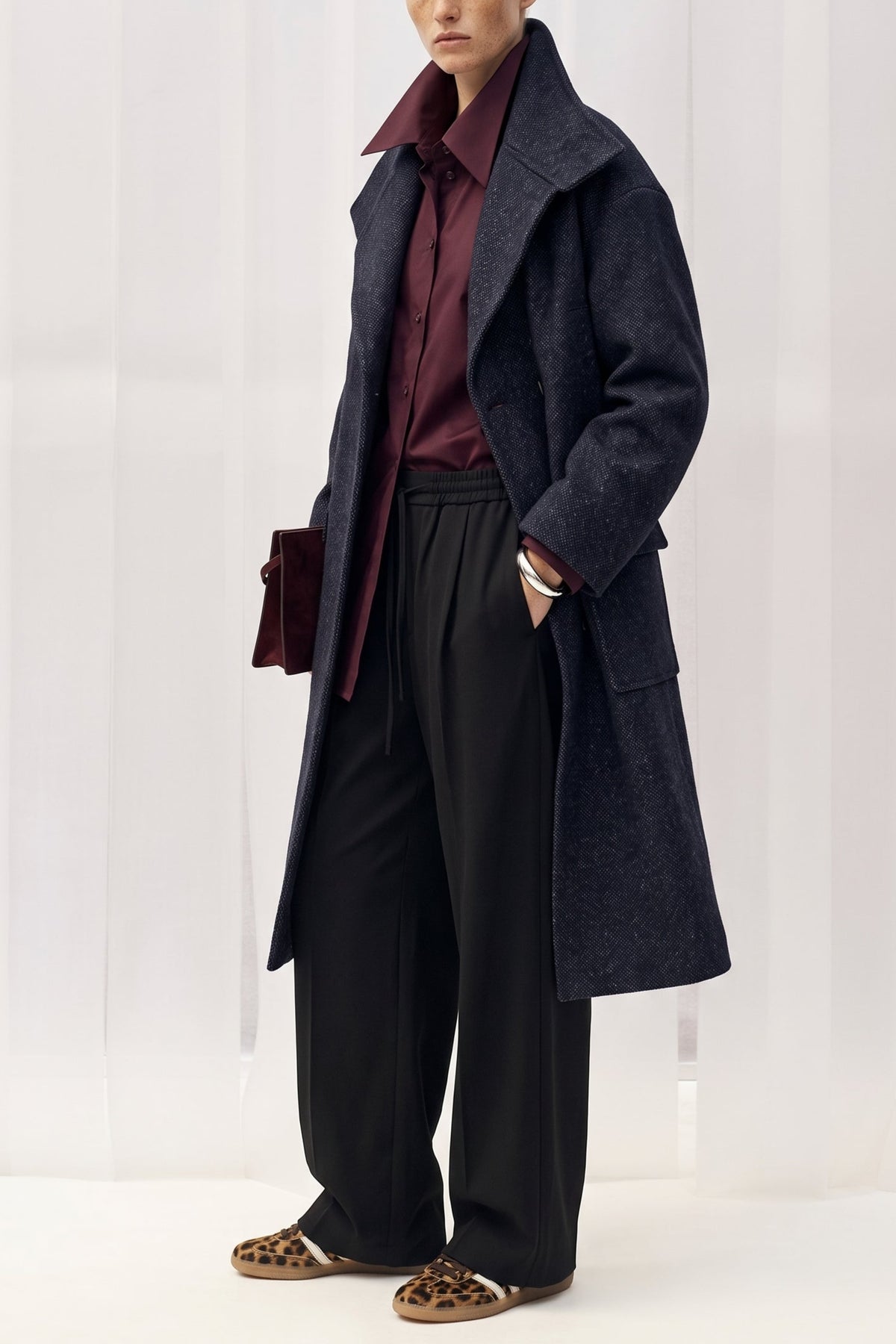 A model stands in a studio wearing a long dark navy wool-blend coat over a burgundy shirt and black wide-leg trousers, with leopard-print loafers and a small dark clutch.
