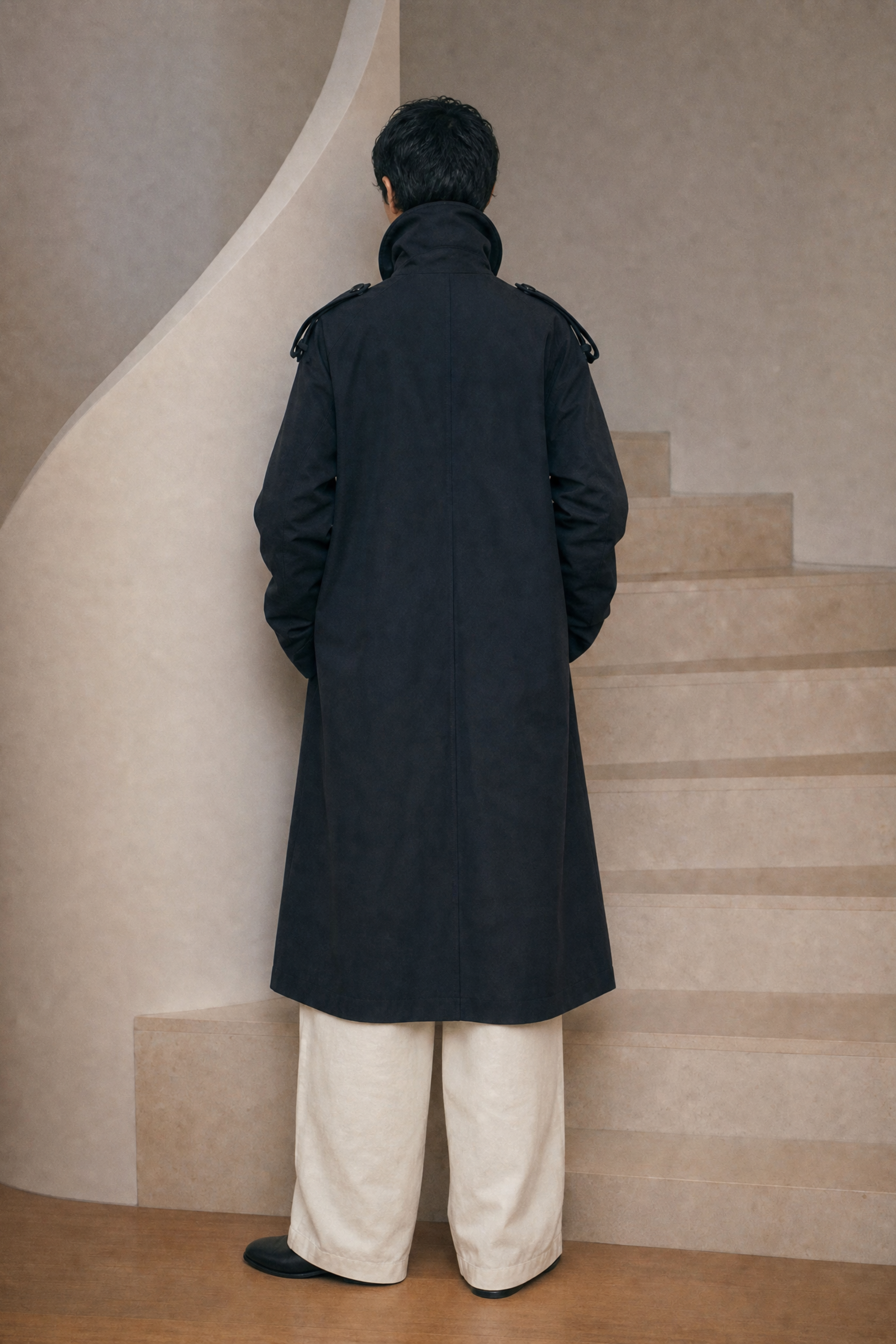 Person wearing a long dark coat standing on a staircase with a neutral wall and floor.