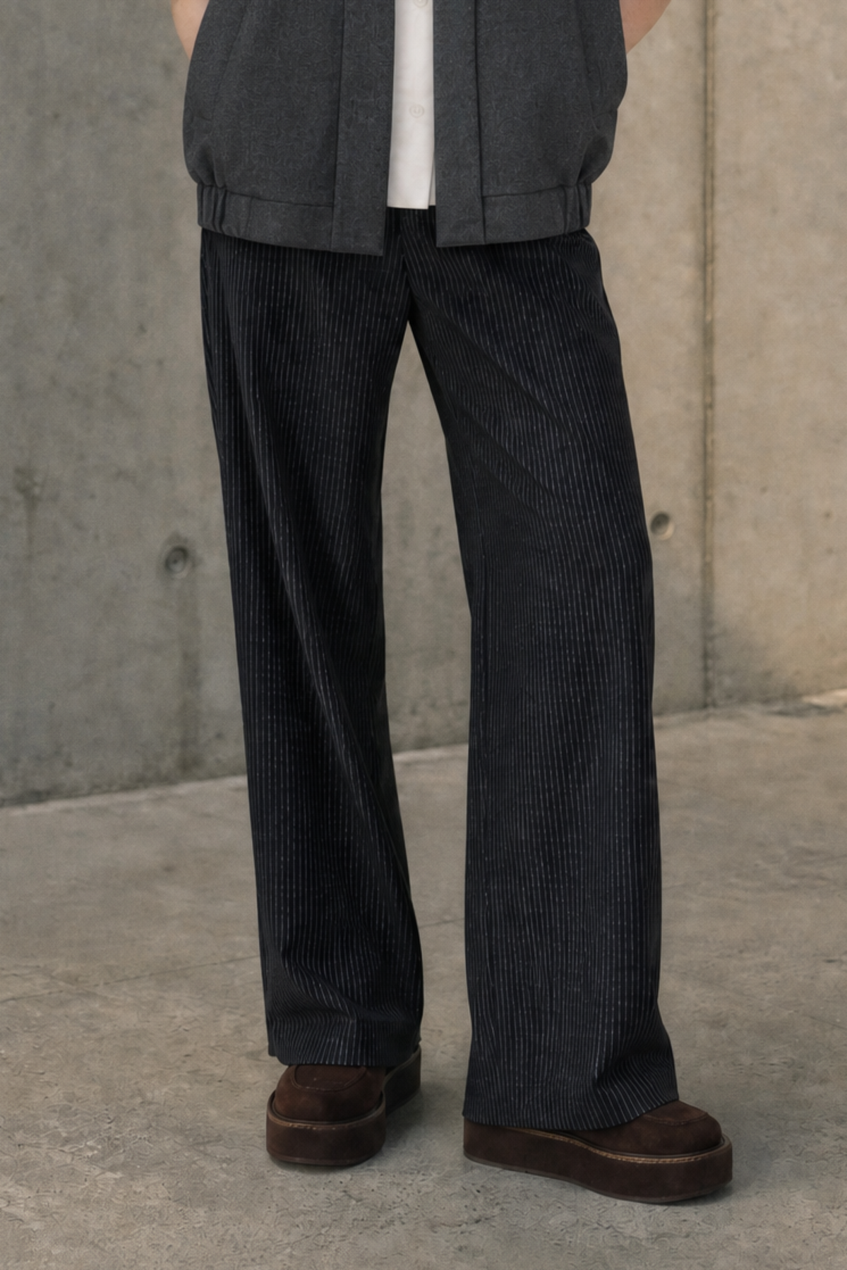 A model stands against a textured concrete wall wearing a dark gray sleeveless vest over a white short-sleeve shirt, paired with dark wide-leg trousers and brown shoes.