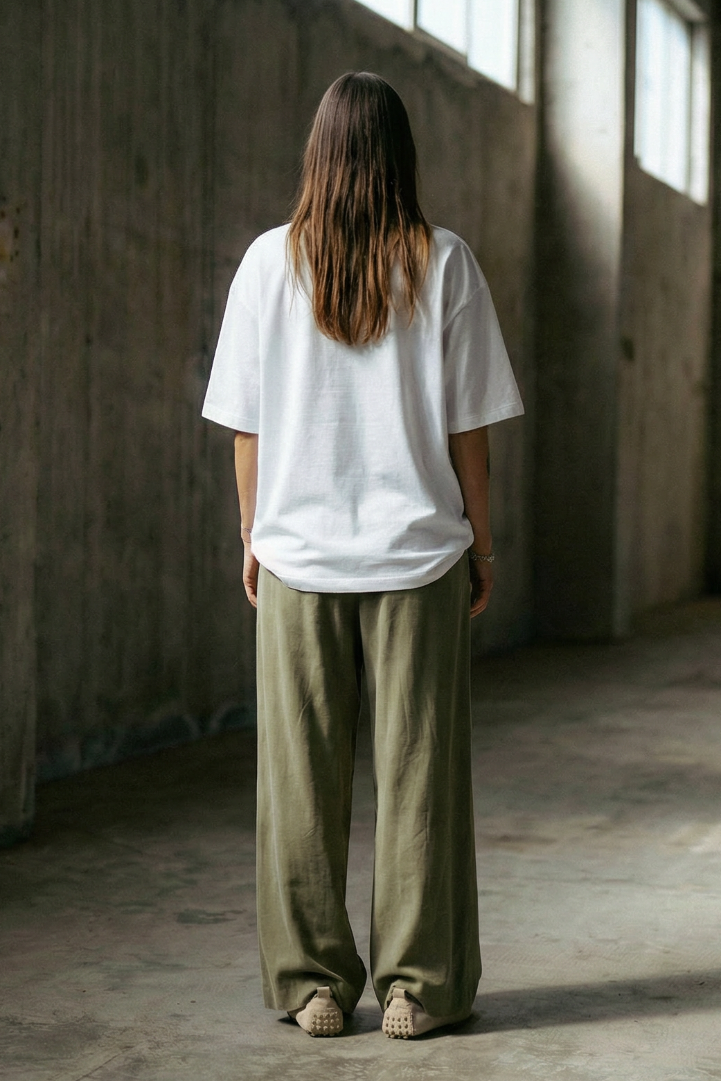 Person wearing a white t-shirt and green pants standing in a sunlit, empty room with concrete walls.
