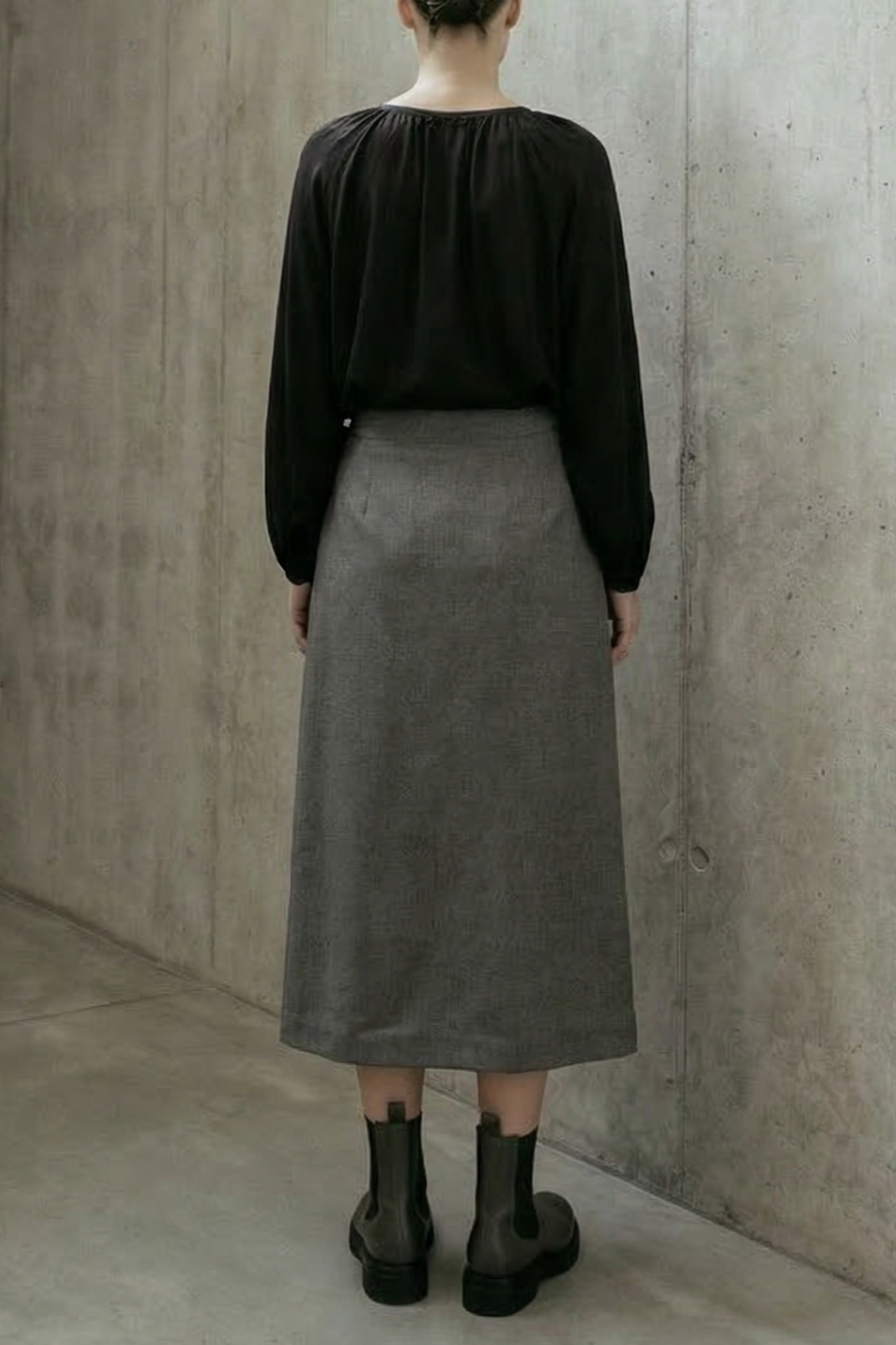 Person wearing a black top and gray skirt standing in a minimalistic concrete room.