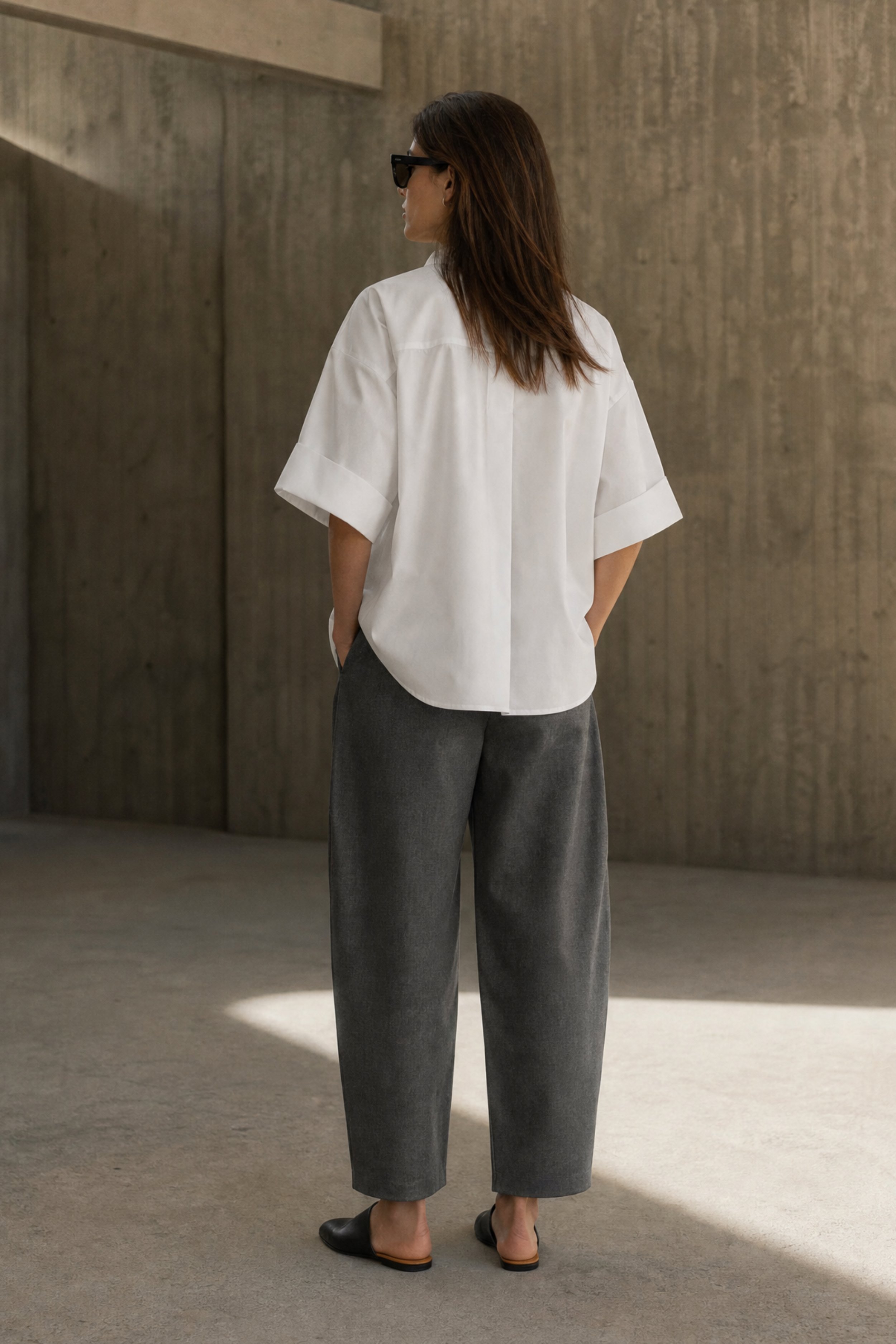 Person wearing a white shirt and gray pants standing in a sunlit room with concrete walls.