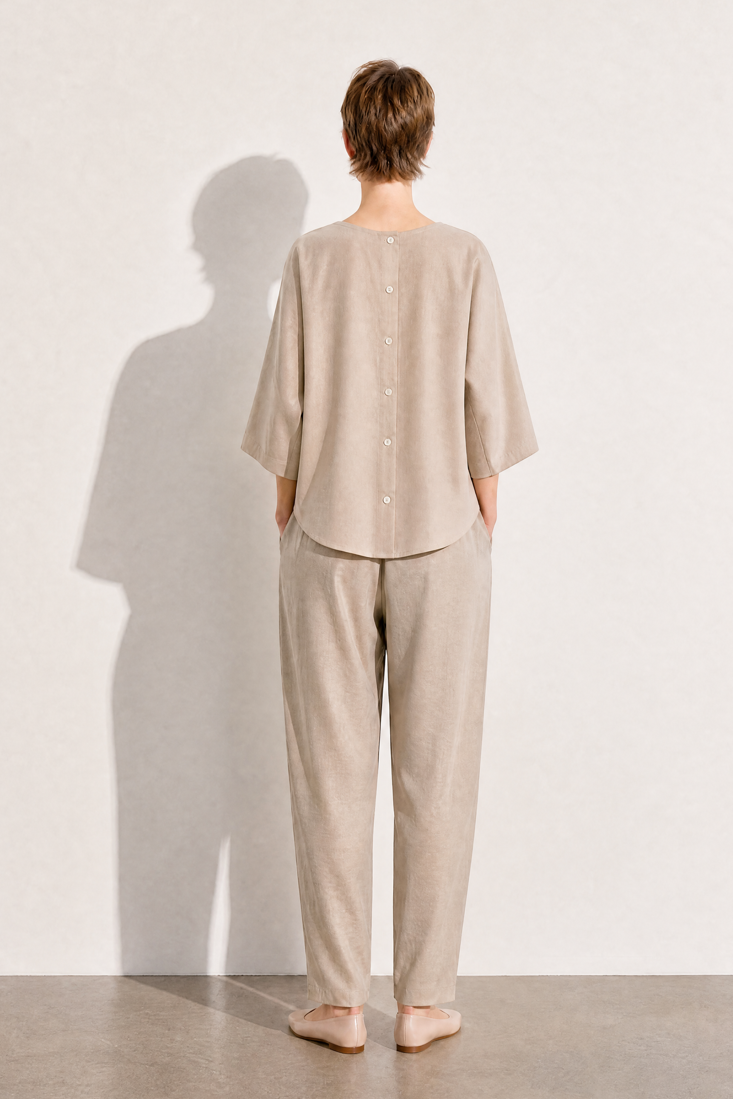 A model stands with their back to the camera wearing a light beige linen two-piece lounge set consisting of a short-sleeve button-up top and matching trousers, set against a minimalist off-white wall with a soft shadow.