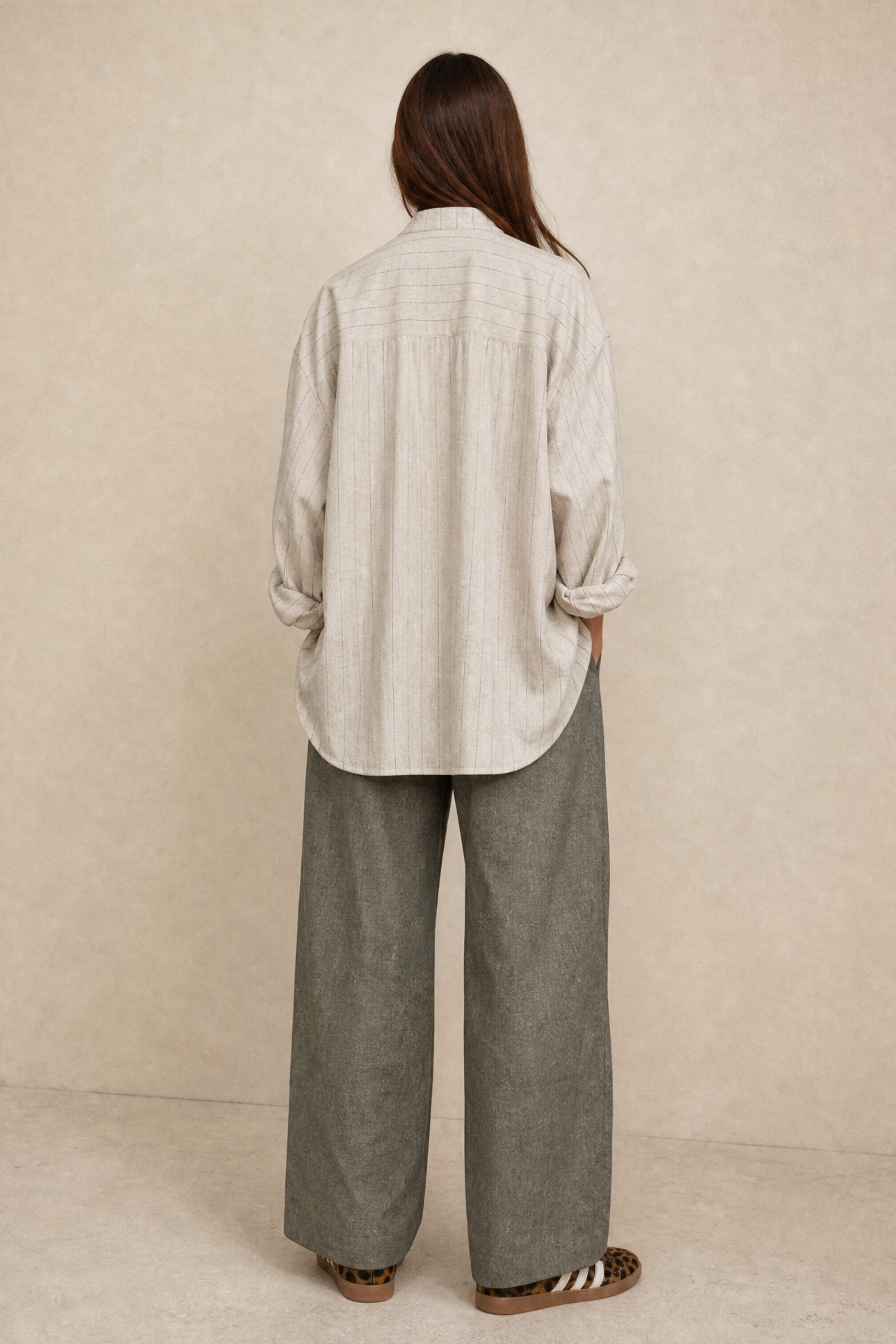 A woman shown from the back wearing a light beige linen shirt with rolled-up sleeves and wide-leg gray trousers, standing against a textured beige backdrop.