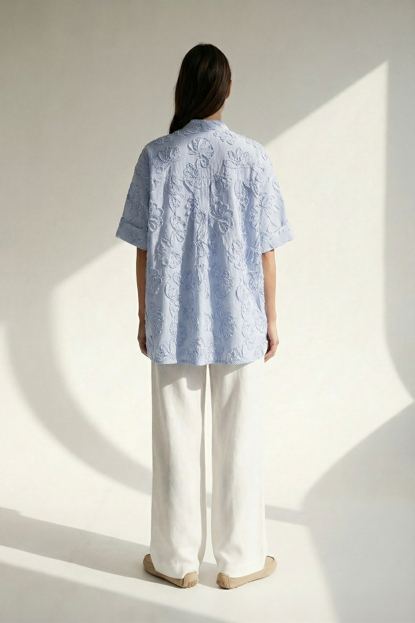 Person wearing a light blue patterned shirt and white pants standing in a well-lit room.