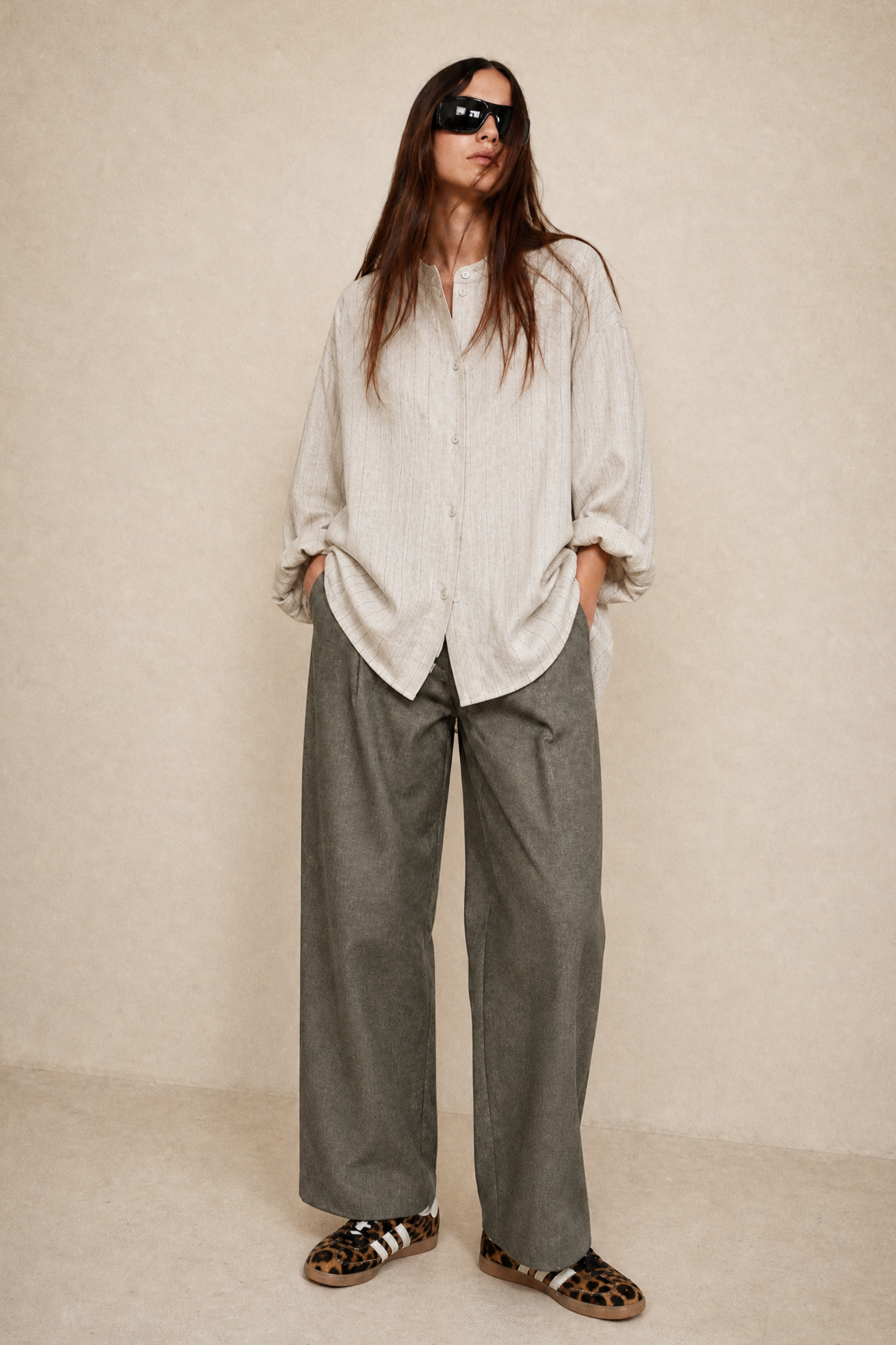 A woman stands against a warm beige studio backdrop wearing a loose off-white linen button-up shirt, wide-leg olive green trousers, and leopard-print slide sandals.