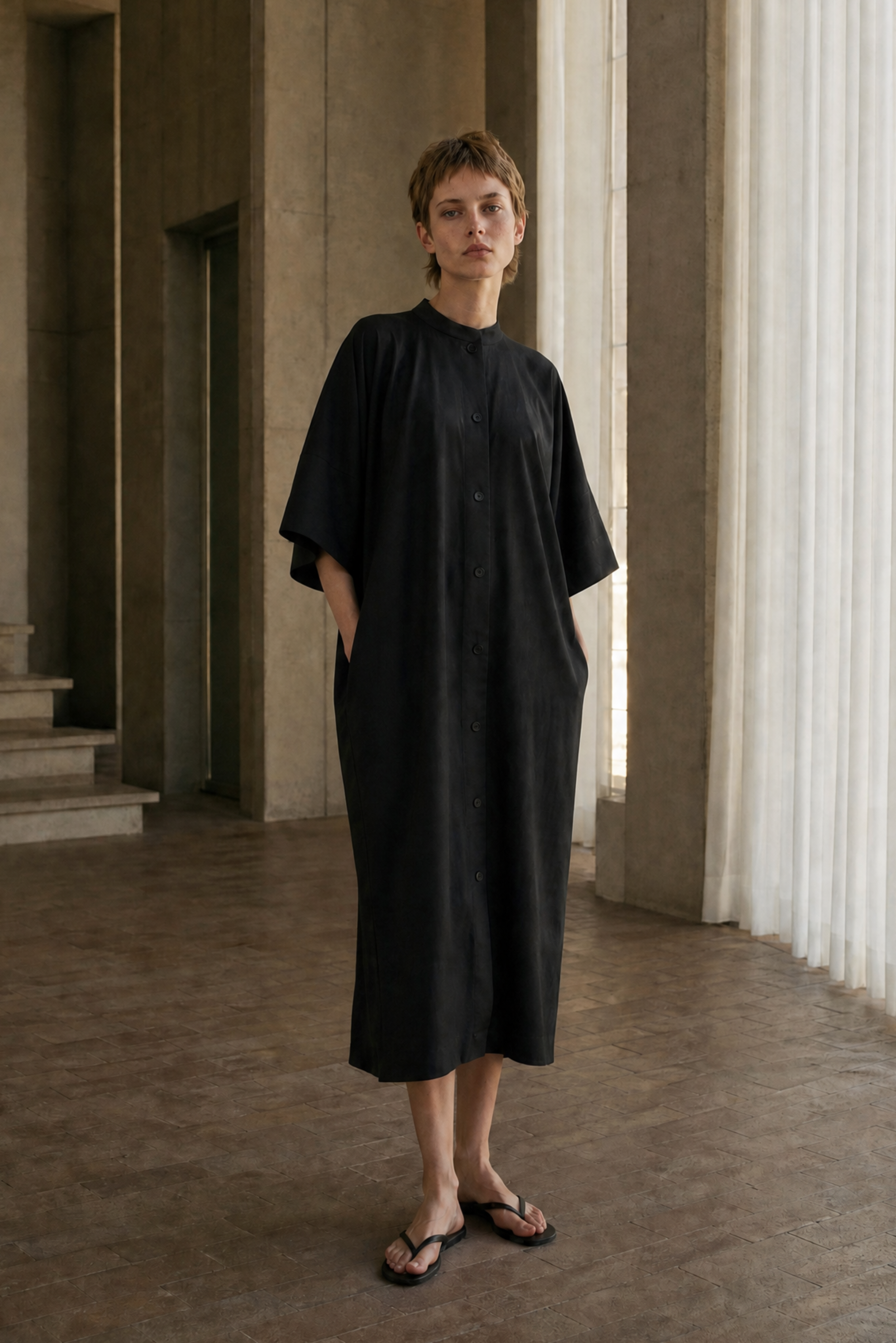 A woman wearing a loose, black midi dress with wide short sleeves and side pockets, standing in a minimalist interior with concrete columns and sheer curtains.